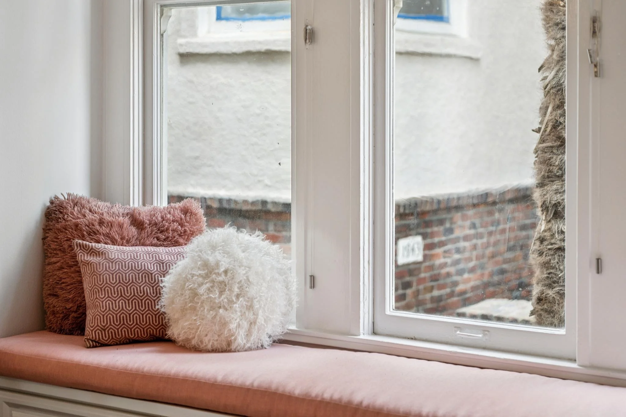 A cozy window seat with pink and textured throw pillows and fluffy white and tan pillows, looking out onto a brick wall and window frame.