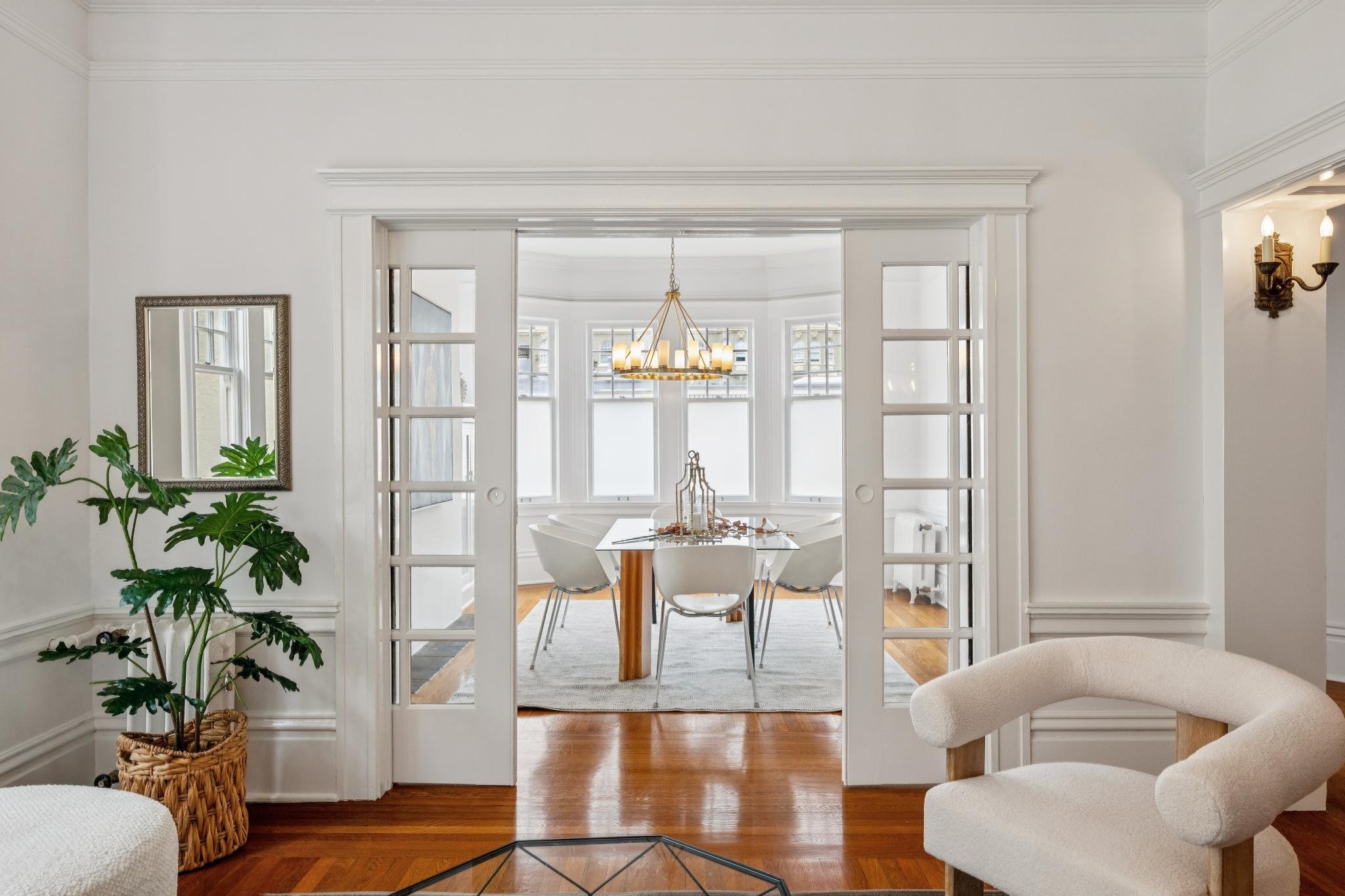 A dining room with a white table and six chairs, three large windows with white frames, a modern chandelier, hardwood floors, and an opening leading to a bright sitting area with a window wall, a wall mirror, and a houseplant in a woven basket.