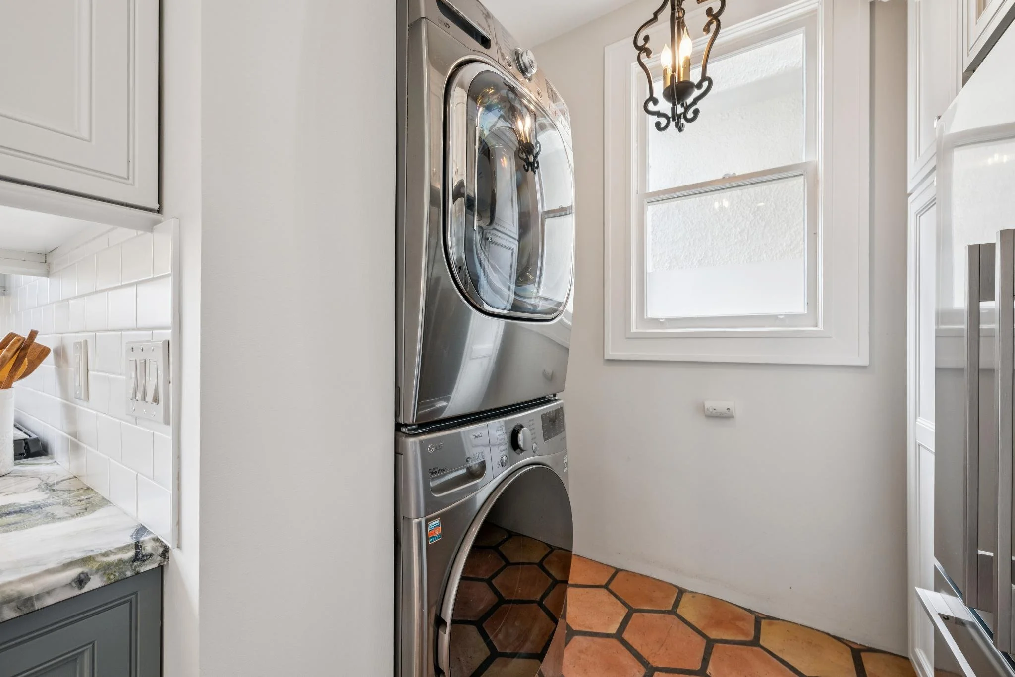 A laundry room with a front-loading washer and dryer stacked next to a window, with tile flooring and white walls.