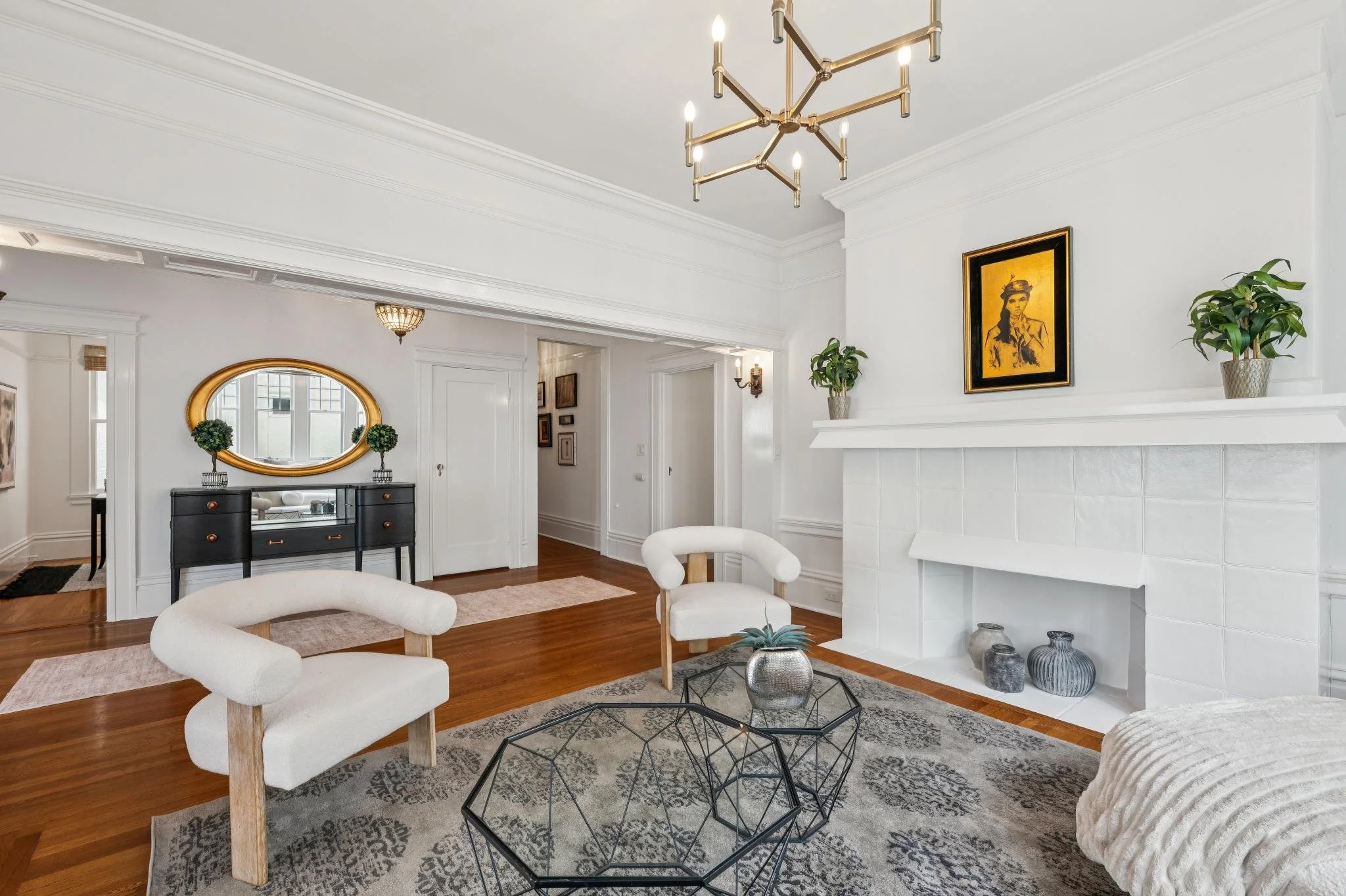 Interior view of a modern living room with white walls, hardwood floors, and decorative crown molding. Contains a white armchair, black sideboard with plants and mirror, framed artwork above fireplace, and metallic geometric coffee table. There are d
