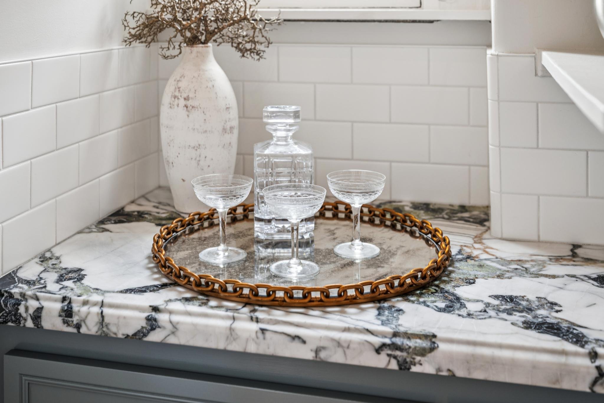 A marble tray with a beaded edge sitting on a marble countertop, holding three crystal coupe glasses and a tall glass water carafe, with a large white vase and dried branches in the background.