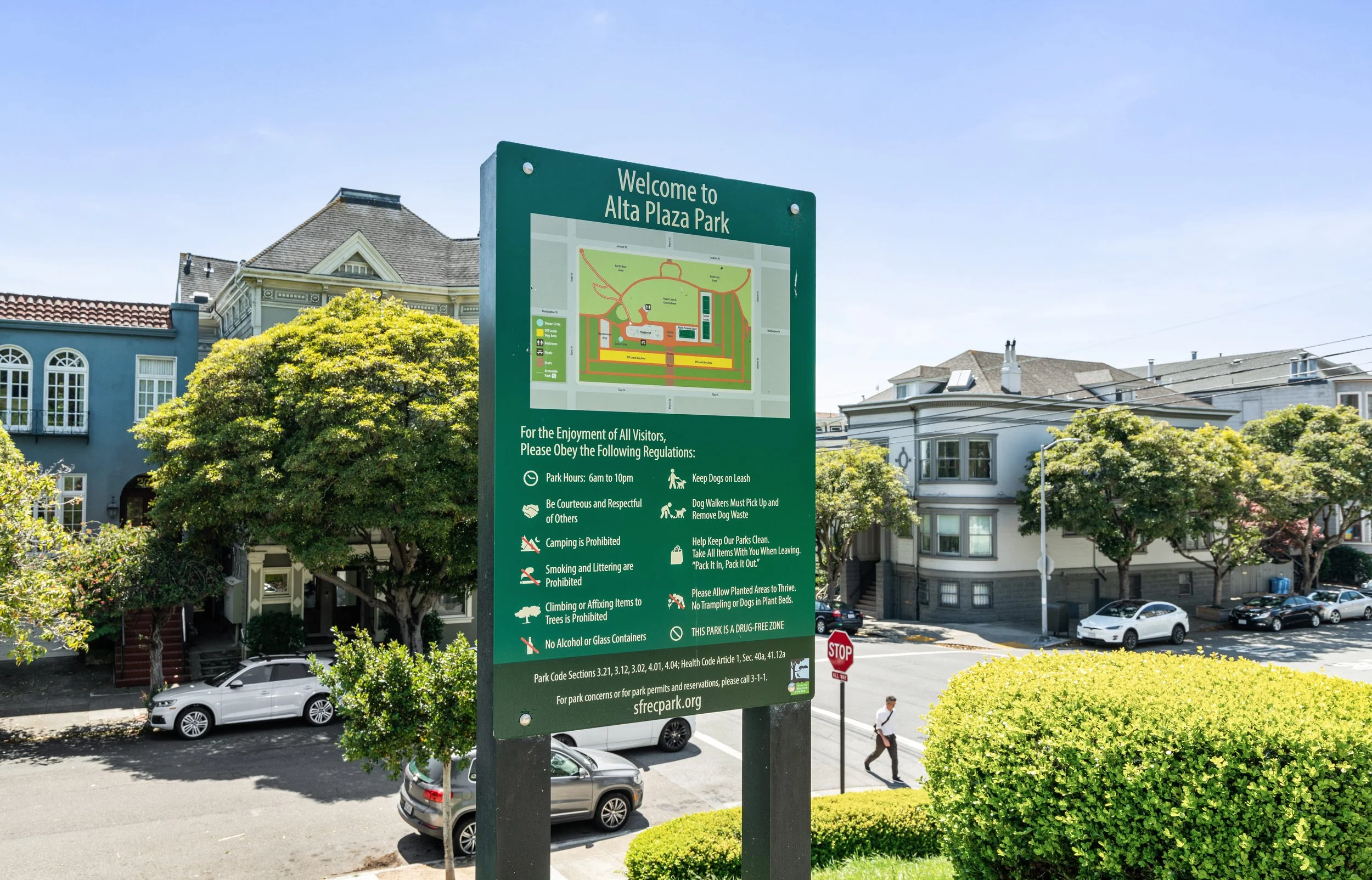 Signboard at Alta Plaza Park indicating park regulations and a map of the park, with residential buildings and parked cars in the background on a sunny day.