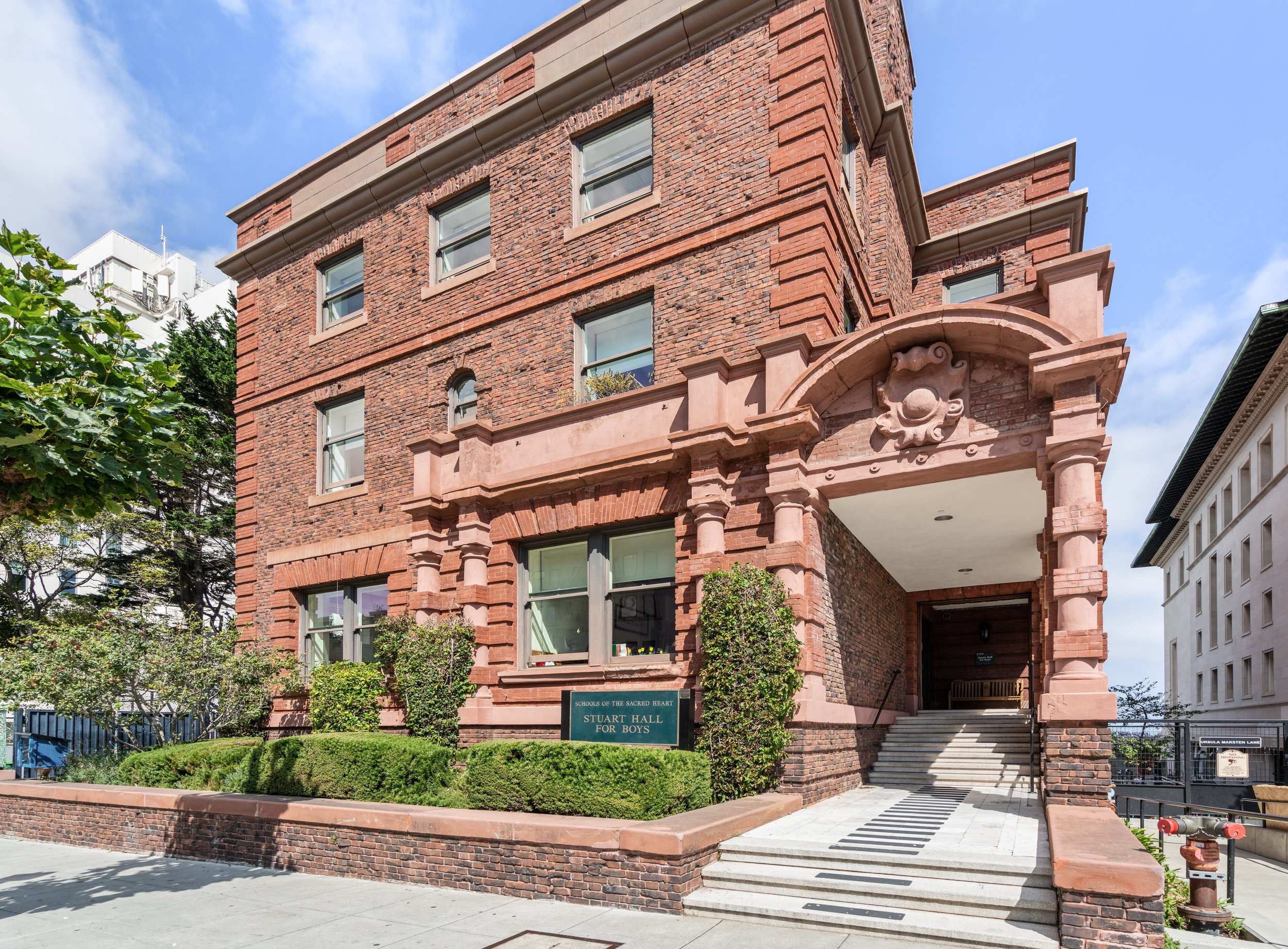 A brick building with a decorative archway and sign that reads 'SCHOOL OF THE SACRED HEART, STUART HALL FOR BOYS.' The building has several windows and is surrounded by greenery, with stairs leading up to the entrance and a clear blue sky above.