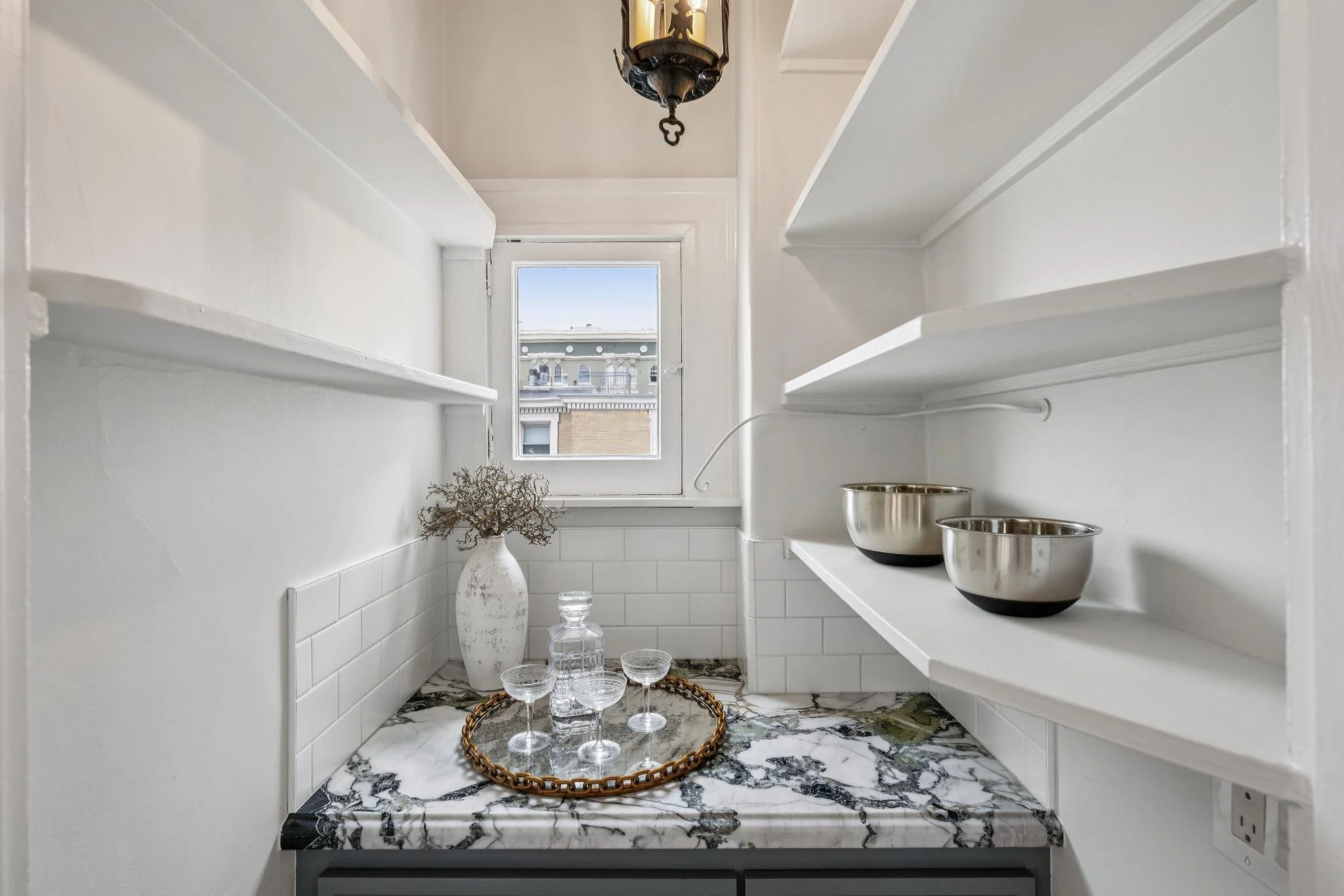 A small kitchen nook with white shelves, a window showing rooftops outside, and a marble countertop with a decorative tray holding glassware, as well as metal bowls and a white vase with dried branches.