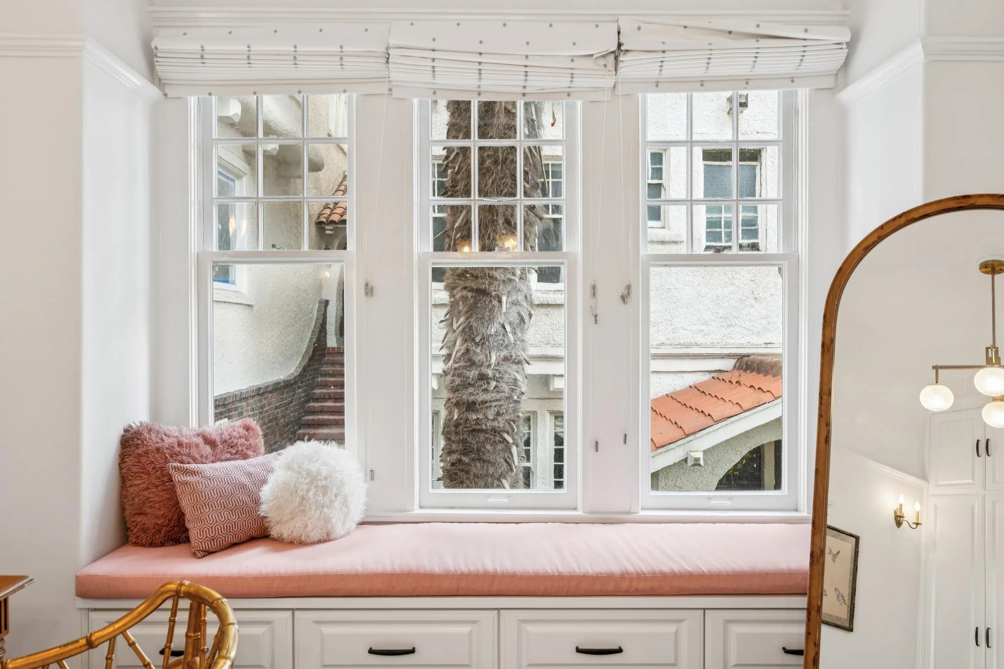A window seat with pink cushion and fluffy white and patterned pillows, overlooking a large palm tree outside, with neighboring white and beige buildings with small windows.