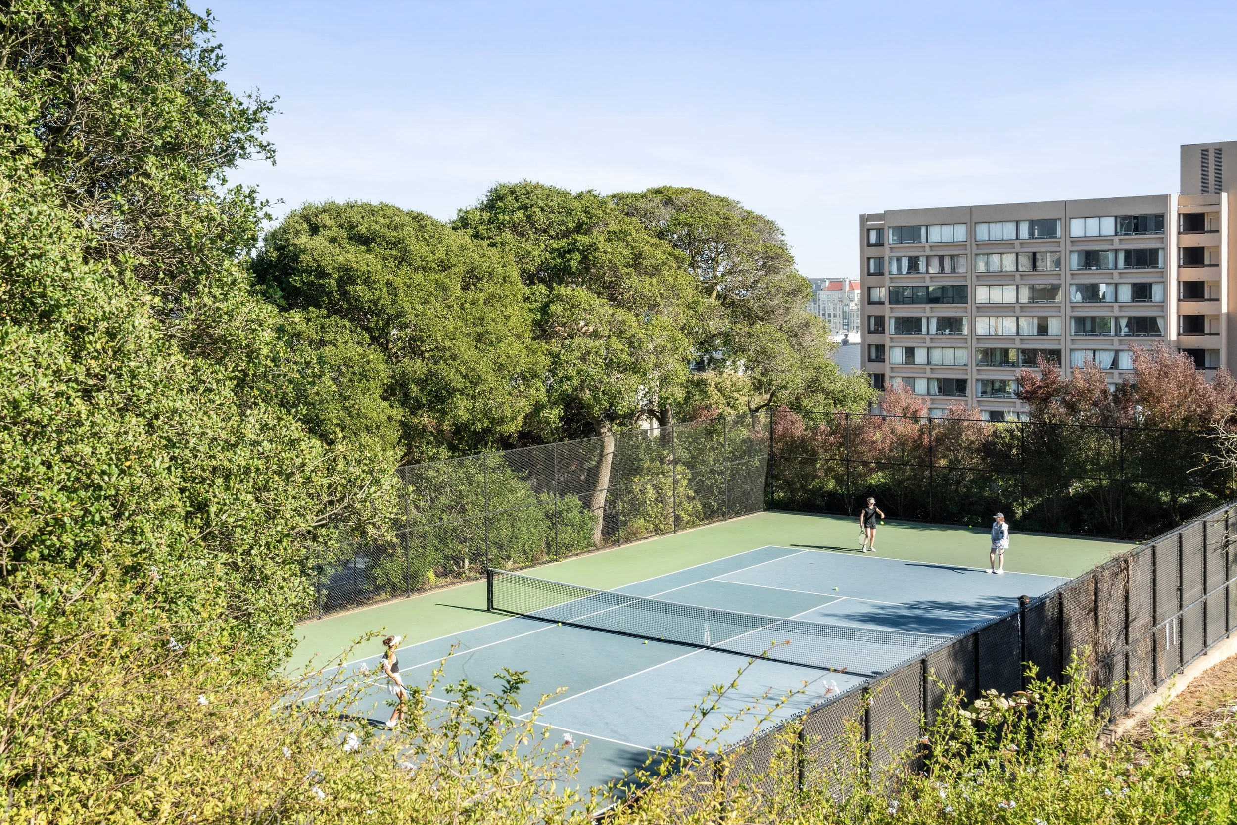 A tennis court surrounded by a black chain-link fence, with three people playing tennis, and large green trees and residential buildings in the background under a clear blue sky.