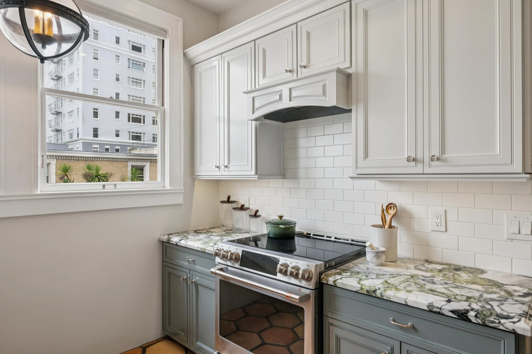 A kitchen with white upper cabinets, a black stove with a stainless steel oven, a marble countertop, and a window showing a cityscape with tall buildings and palm trees outside.