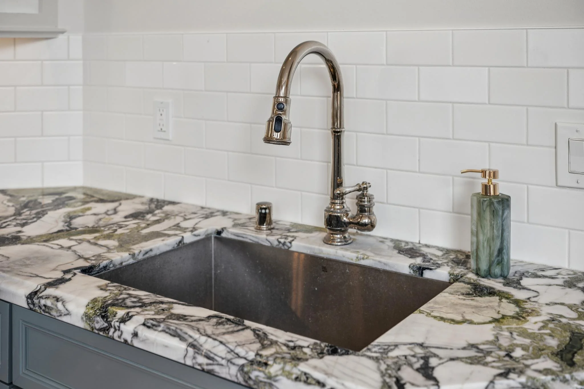 Kitchen sink with a marble countertop and a chrome faucet, soap dispenser, and white subway tile backsplash.