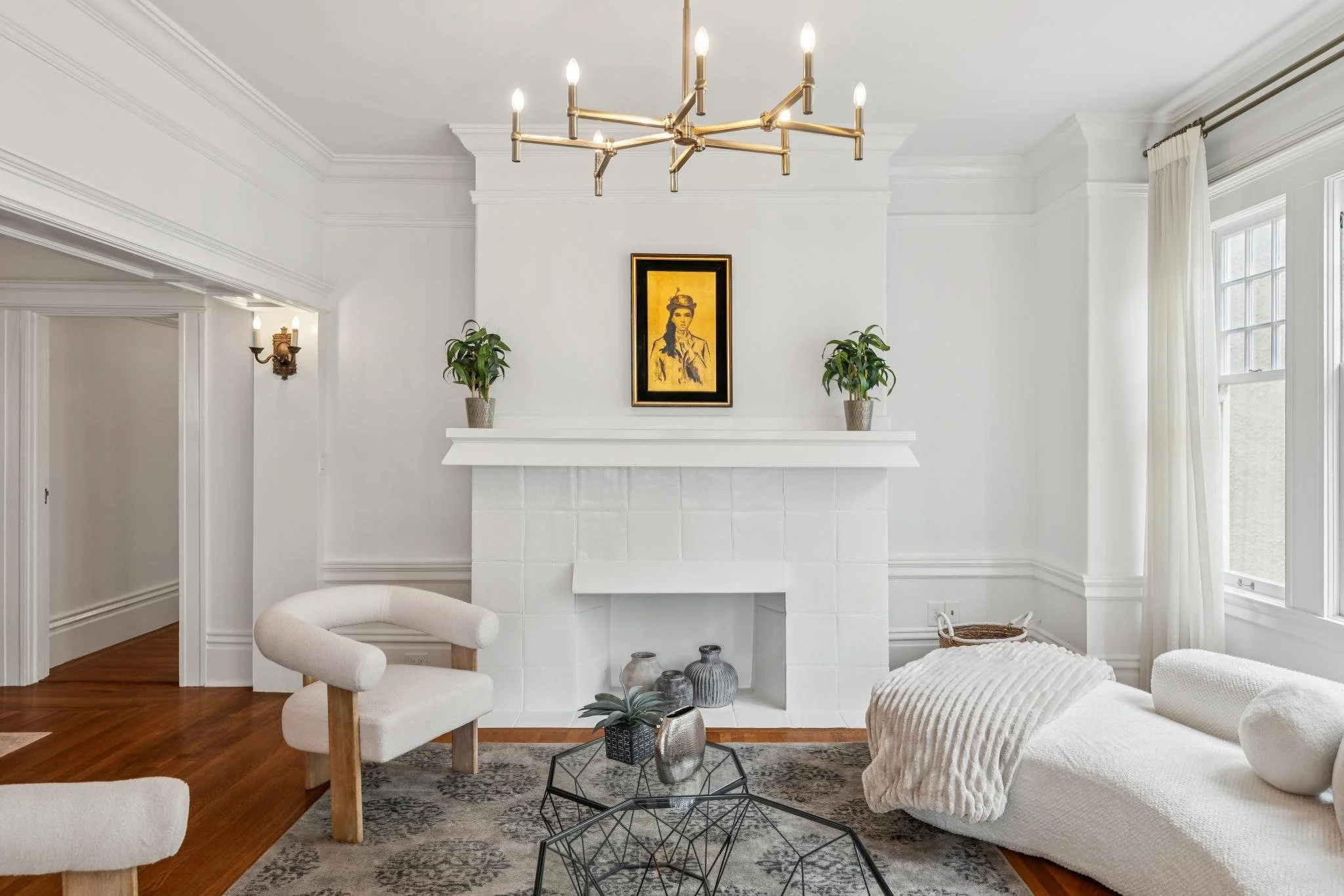 A living room with white walls and high ceilings featuring crown molding, with a modern gold chandelier hanging from the ceiling. There is a white fireplace with a framed yellow artwork above it, flanked by two potted plants. The room has floor-to-ce