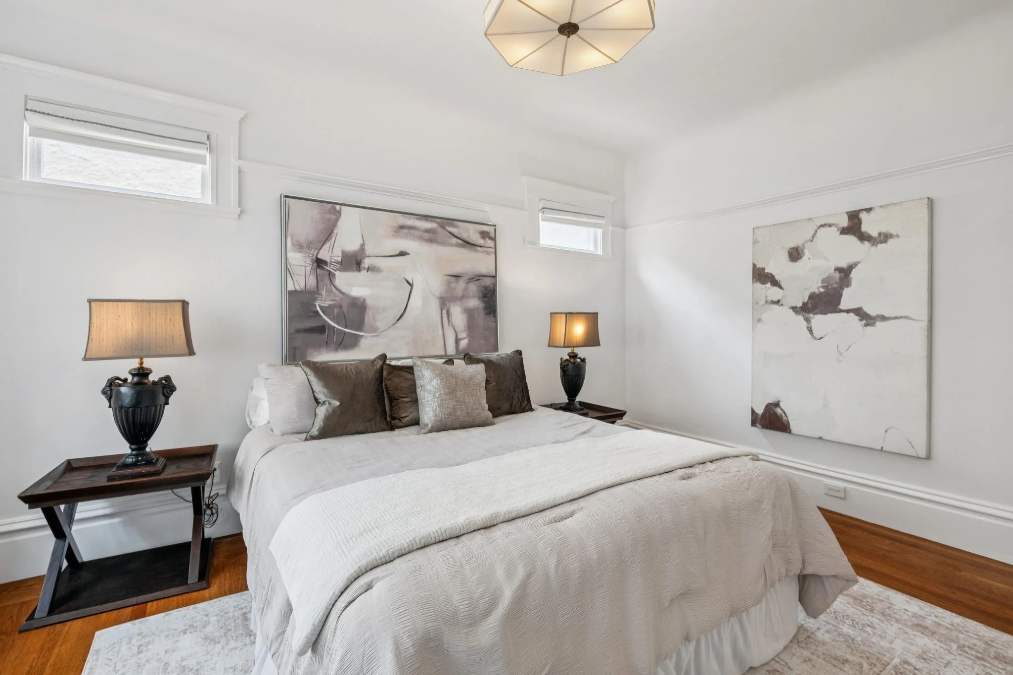 A bedroom with a large bed covered in white bedding, topped with multiple pillows in shades of brown and beige. There are two black bedside lamps with beige lampshades on wooden nightstands beside the bed. The walls are white with decorative molding,