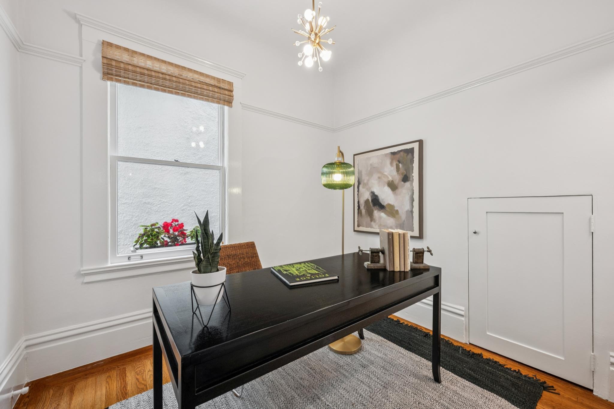 A modern, minimalist home office with a black desk, decorative items, plants, a window with a bamboo shade, a white wall with artwork, a ceiling light fixture, and a small door.