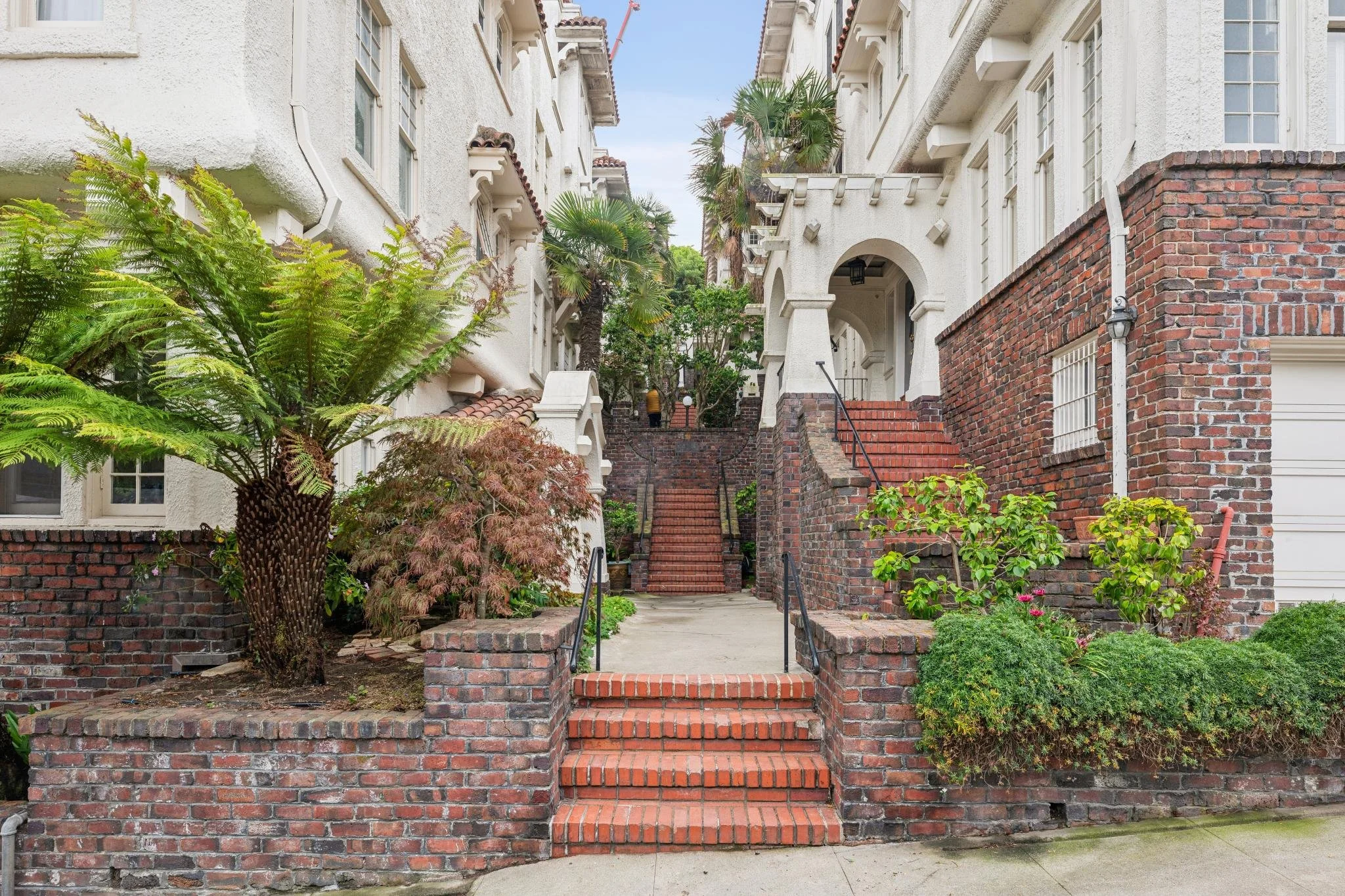 A pathway leading up stairs between two white stucco apartment buildings with red brick accents, lush greenery, and potted plants.