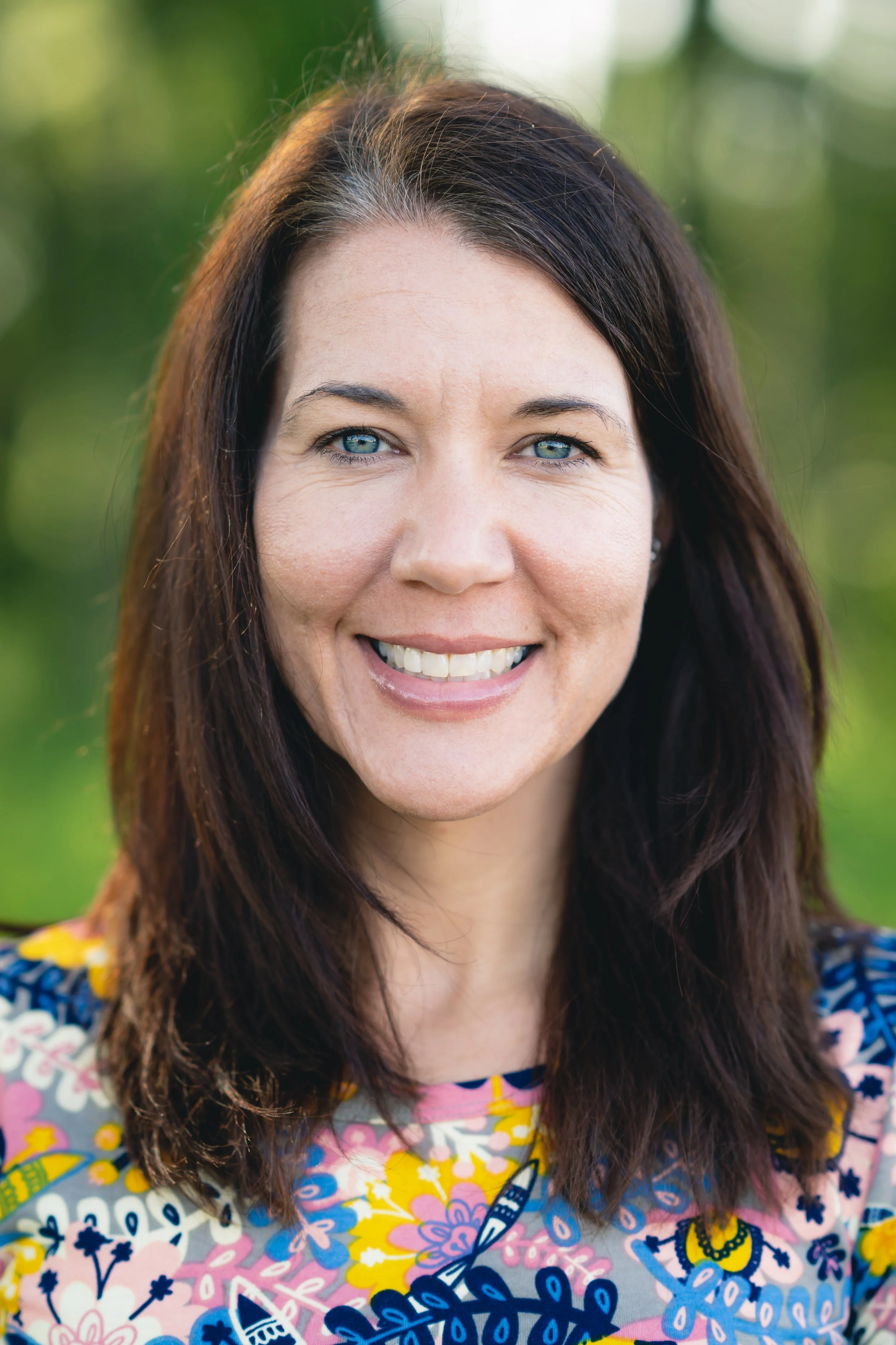 Close-up of a female doctor with long dark brown hair, blue eyes, and a big smile, wearing a colorful, patterned top, outdoors with blurred green foliage in the background.
