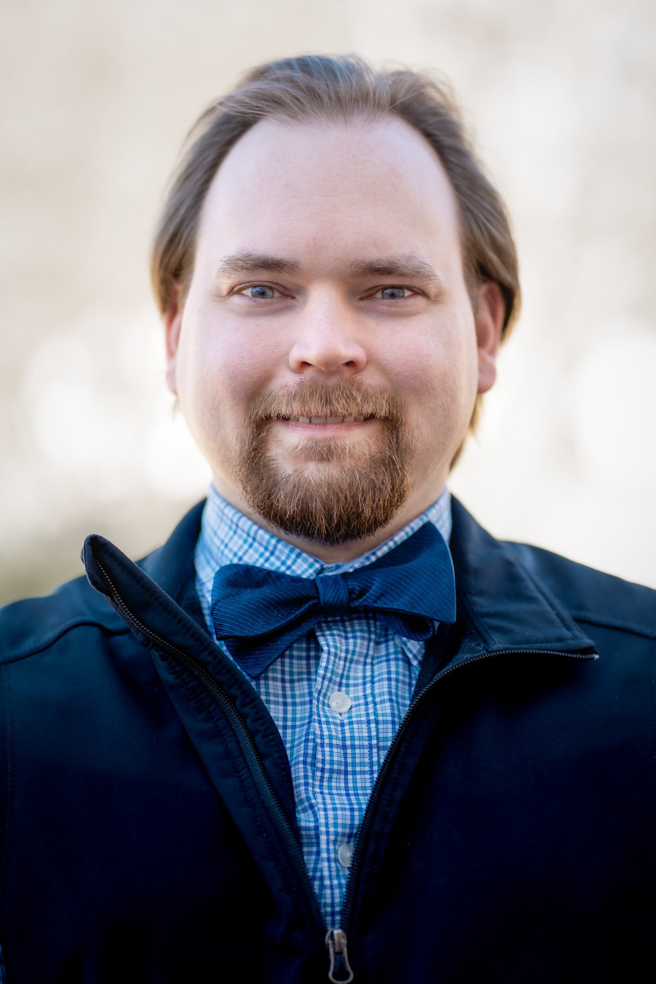 Close-up photo of a smiling man with blue eyes, brown hair, and a beard, wearing a blue checked shirt and a navy jacket with a zipper, outdoors in a natural setting.