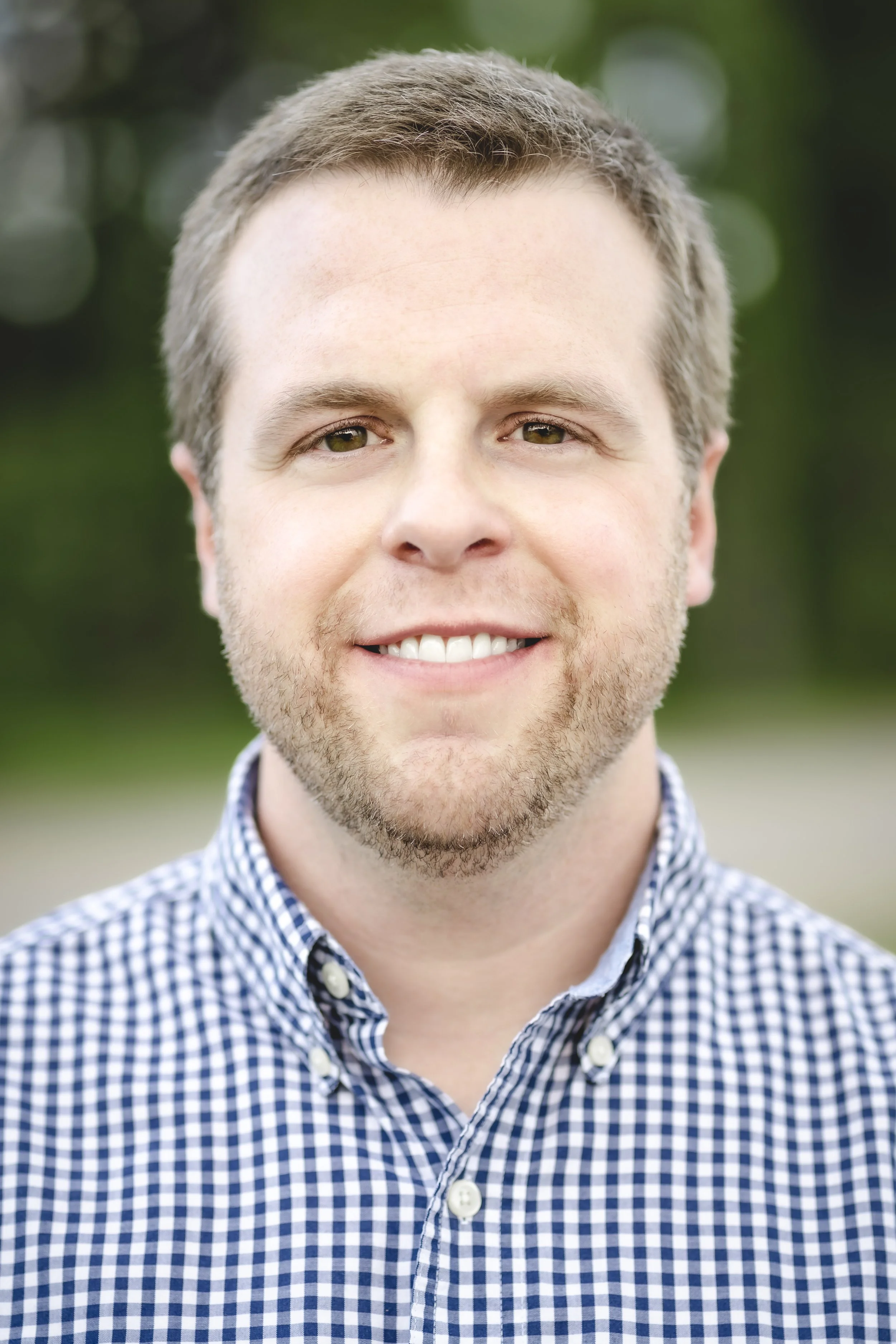 Close-up of a smiling man with brown hair, hazel eyes, and light facial hair, wearing a blue and white checkered shirt, outdoors with a blurred green background.