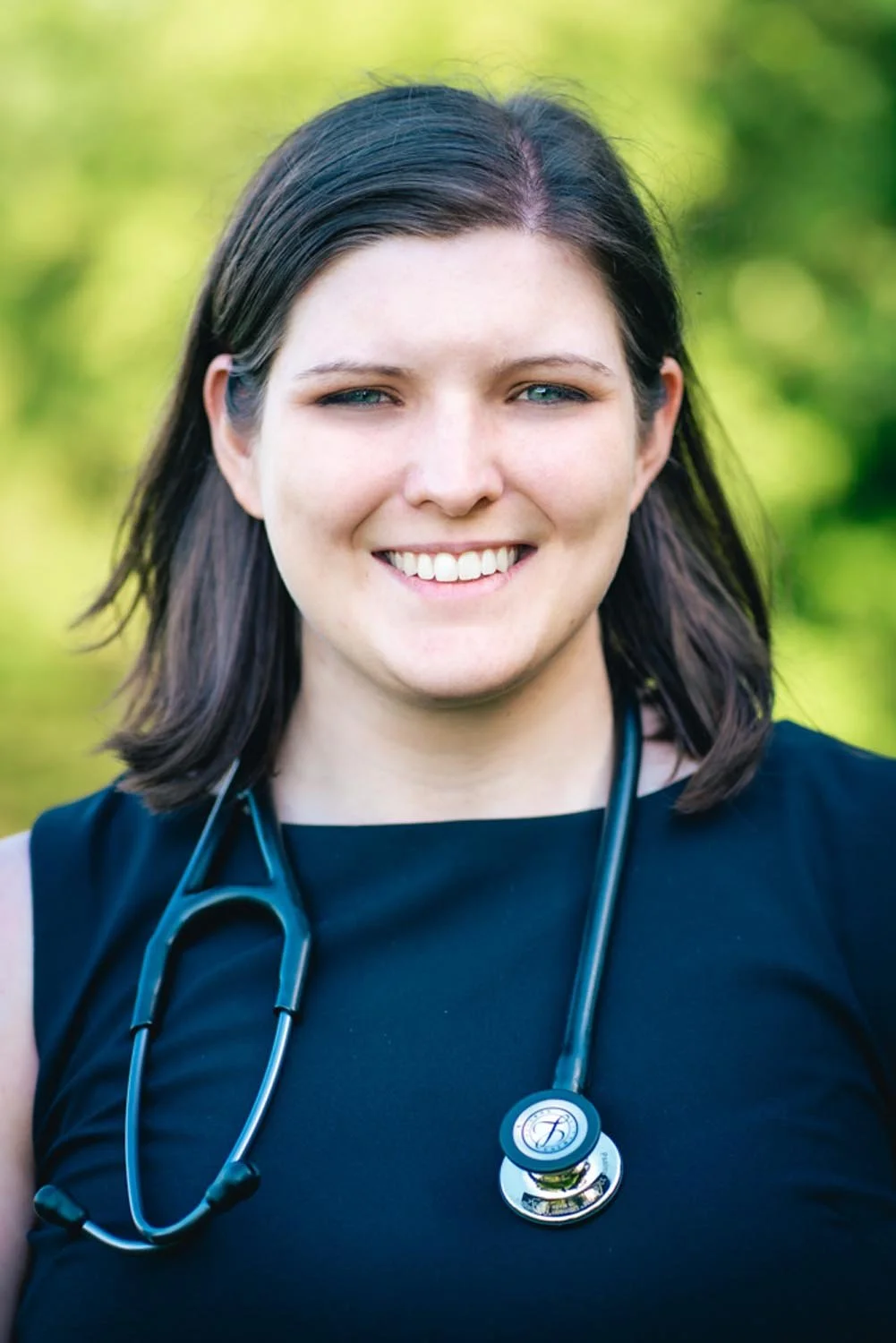 A smiling woman with dark brown hair and blue eyes wearing a black top and a stethoscope around her neck, outdoors with blurred green trees in the background.