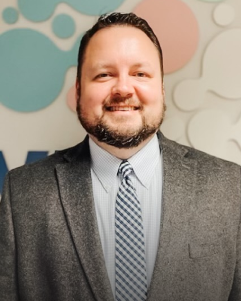 A smiling man in a gray suit with a checkered tie, standing in front of a colorful abstract background.