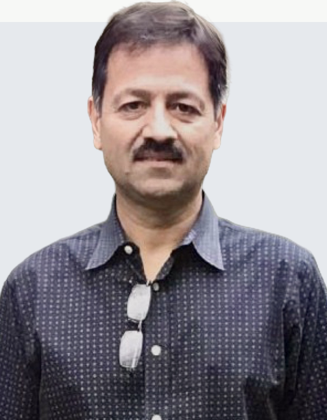 A man with dark hair and a mustache wearing a dark patterned button-up shirt, standing against a light gray background.