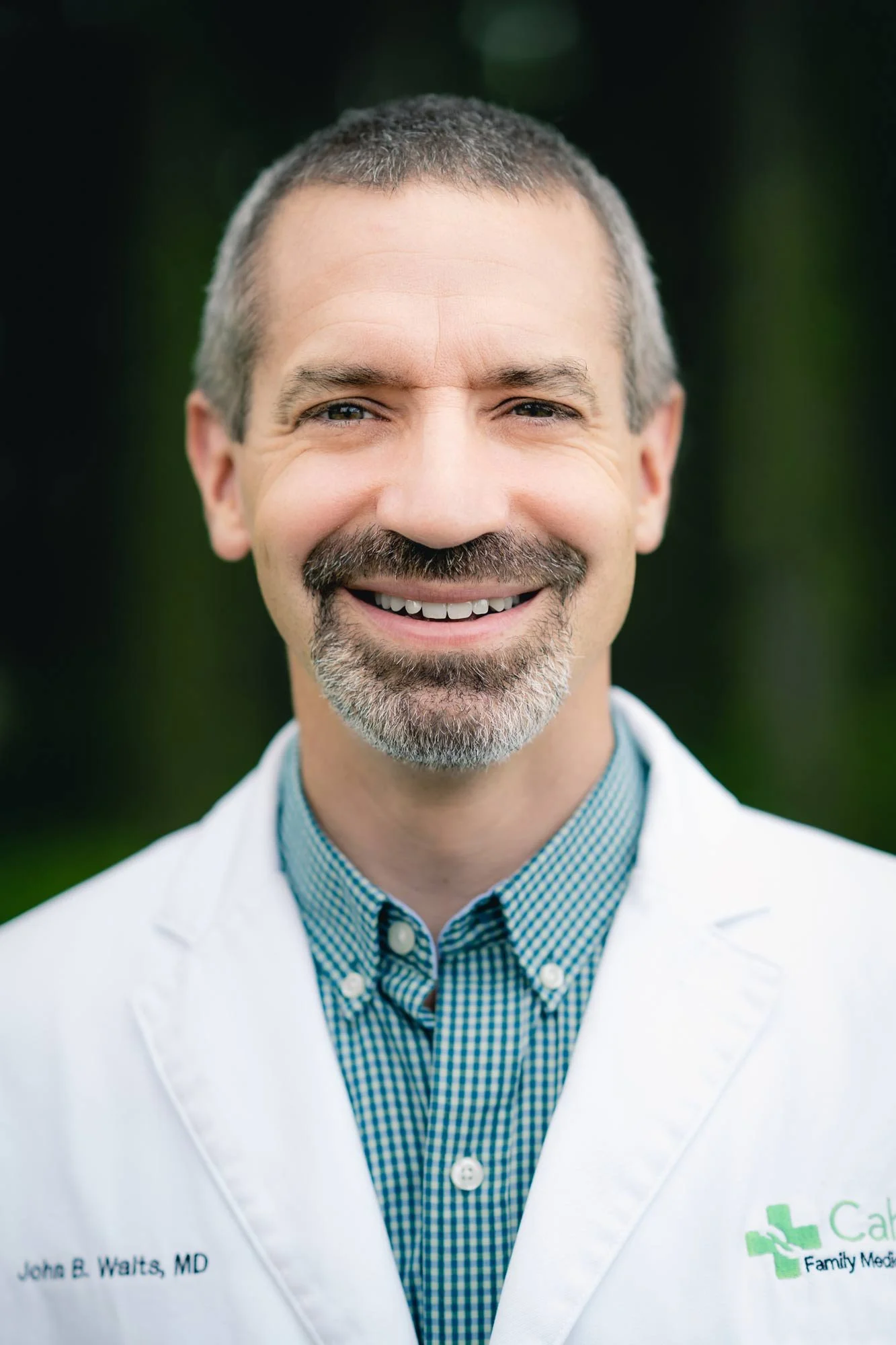 Close-up portrait of a John B. Waits, MD with short dark hair and a beard, smiling, wearing a white medical coat over a blue checkered shirt, standing outdoors with green blurred background.