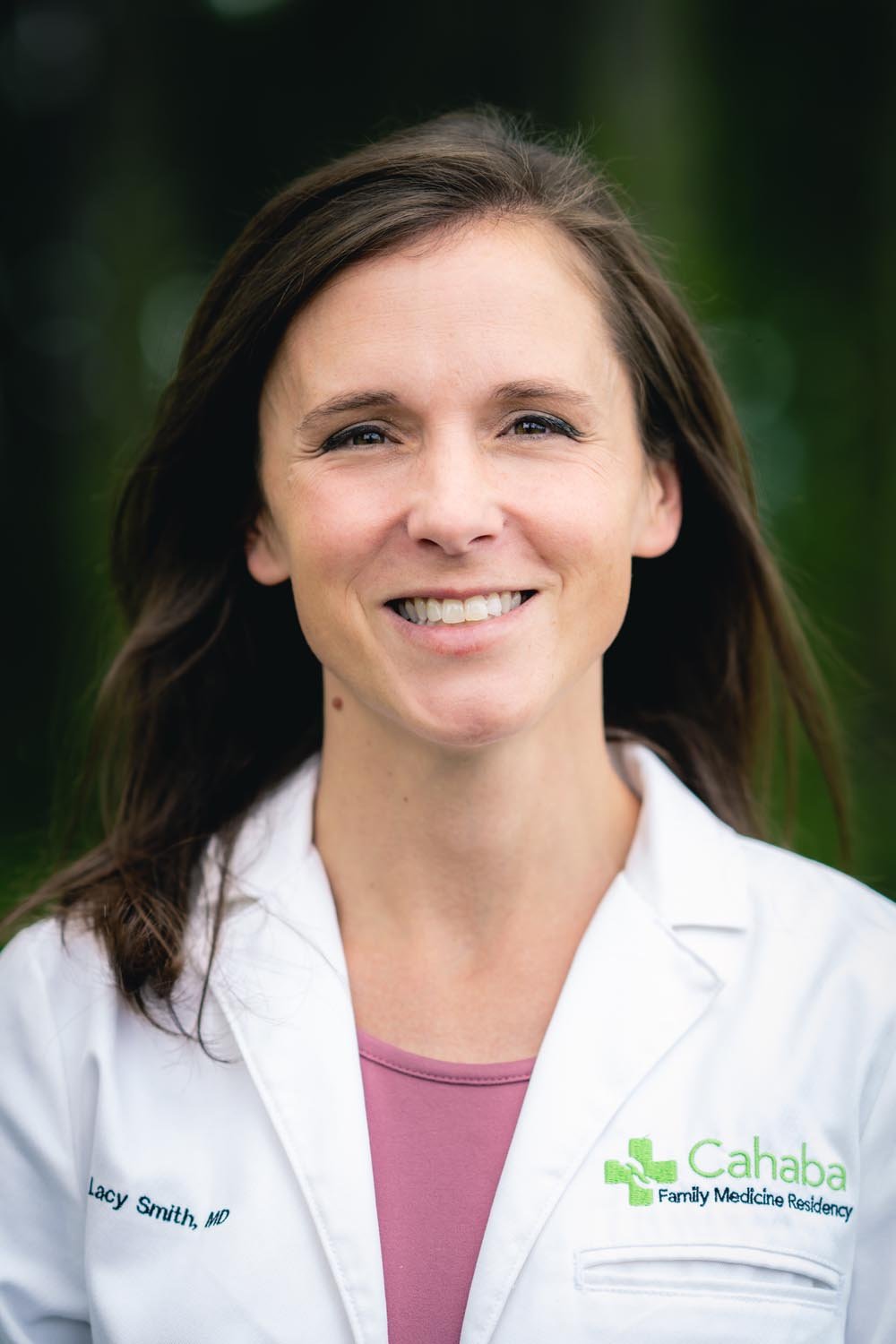 A smiling Lacy Smith, MD, wearing a white medical coat with the logo 'Cahaba Family Medicine Residency' and the name tag 'Lacy Smith, MD,' standing outdoors with a blurred green background.