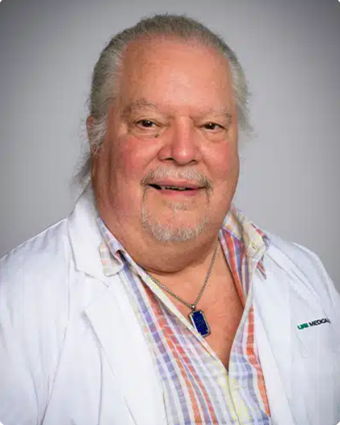 A middle-aged man with gray hair and a beard, wearing a white lab coat over a colorful plaid shirt, and a blue pendant necklace, smiling at the camera.