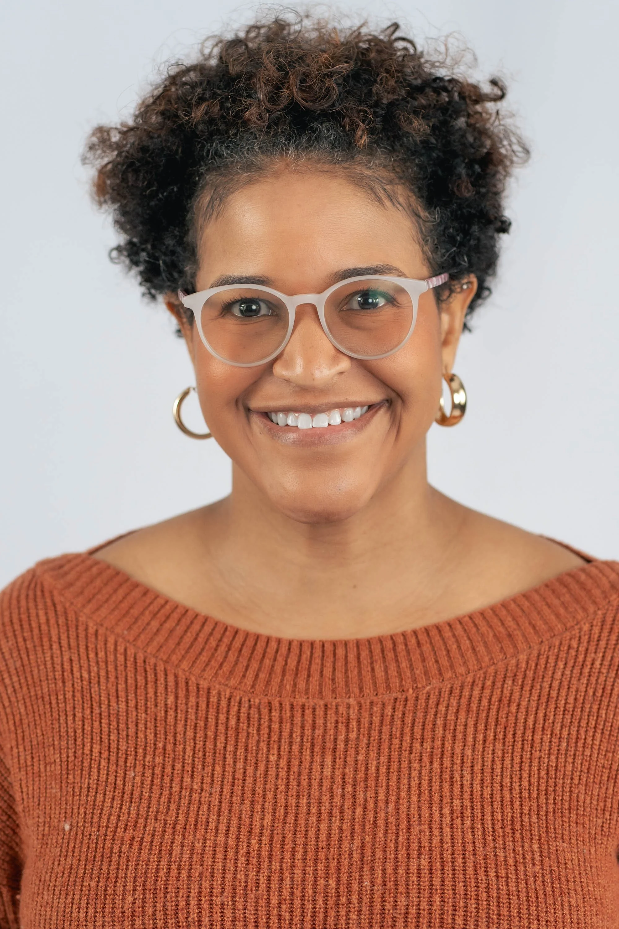 A smiling woman with curly hair, wearing glasses, hoop earrings, and an orange ruffled sweater.