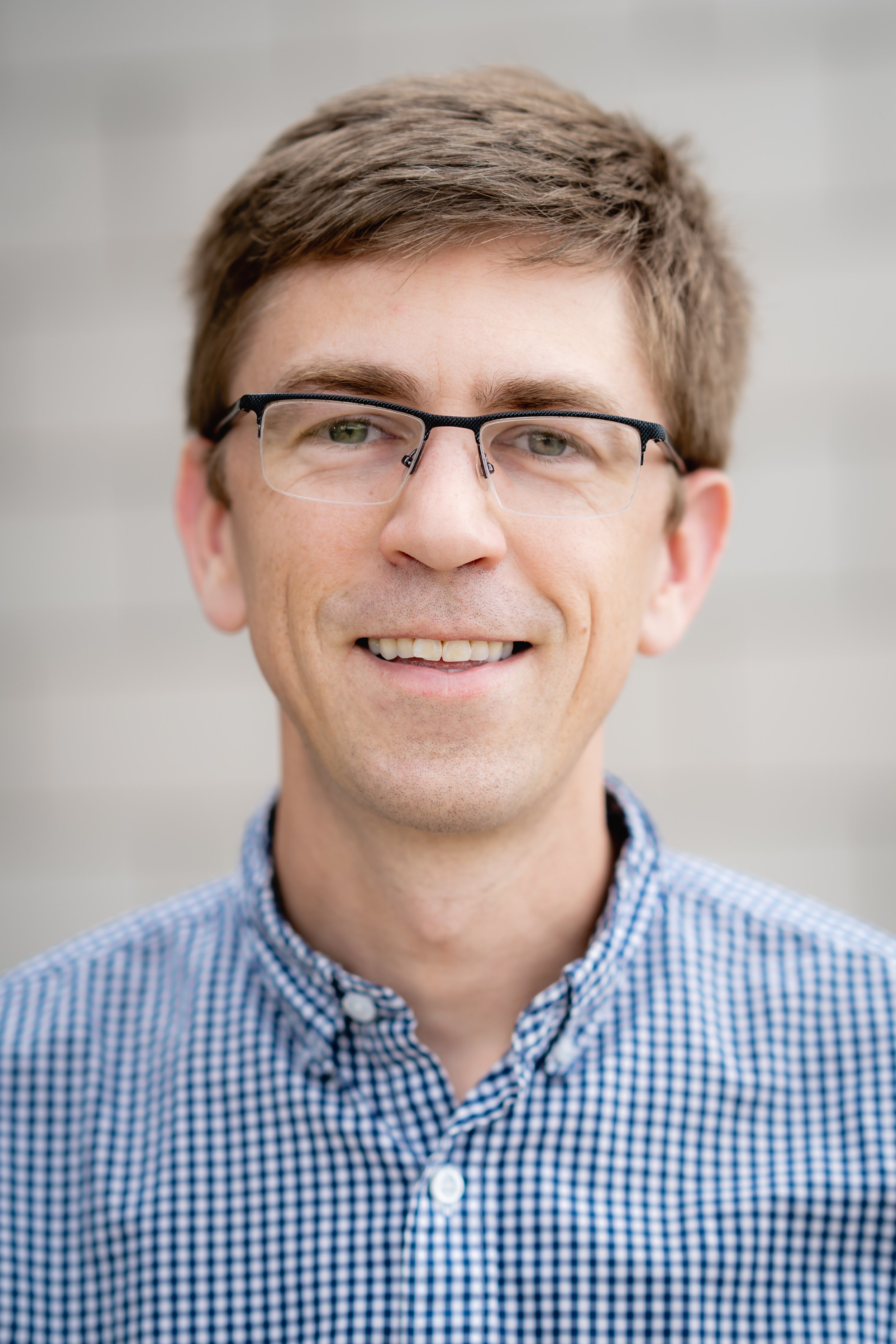 Close-up of a young man with short light brown hair, wearing glasses and a blue checkered shirt, smiling in front of a plain background.