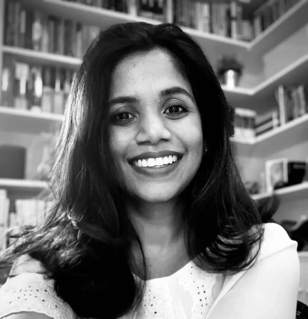 A smiling woman with long dark hair in a room with bookshelves in the background.