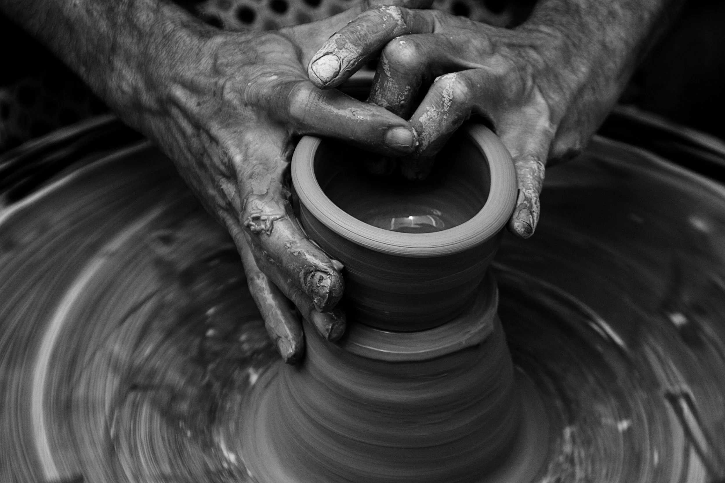 A person shaping pottery on a spinning pottery wheel with hands covered in clay, in black and white.