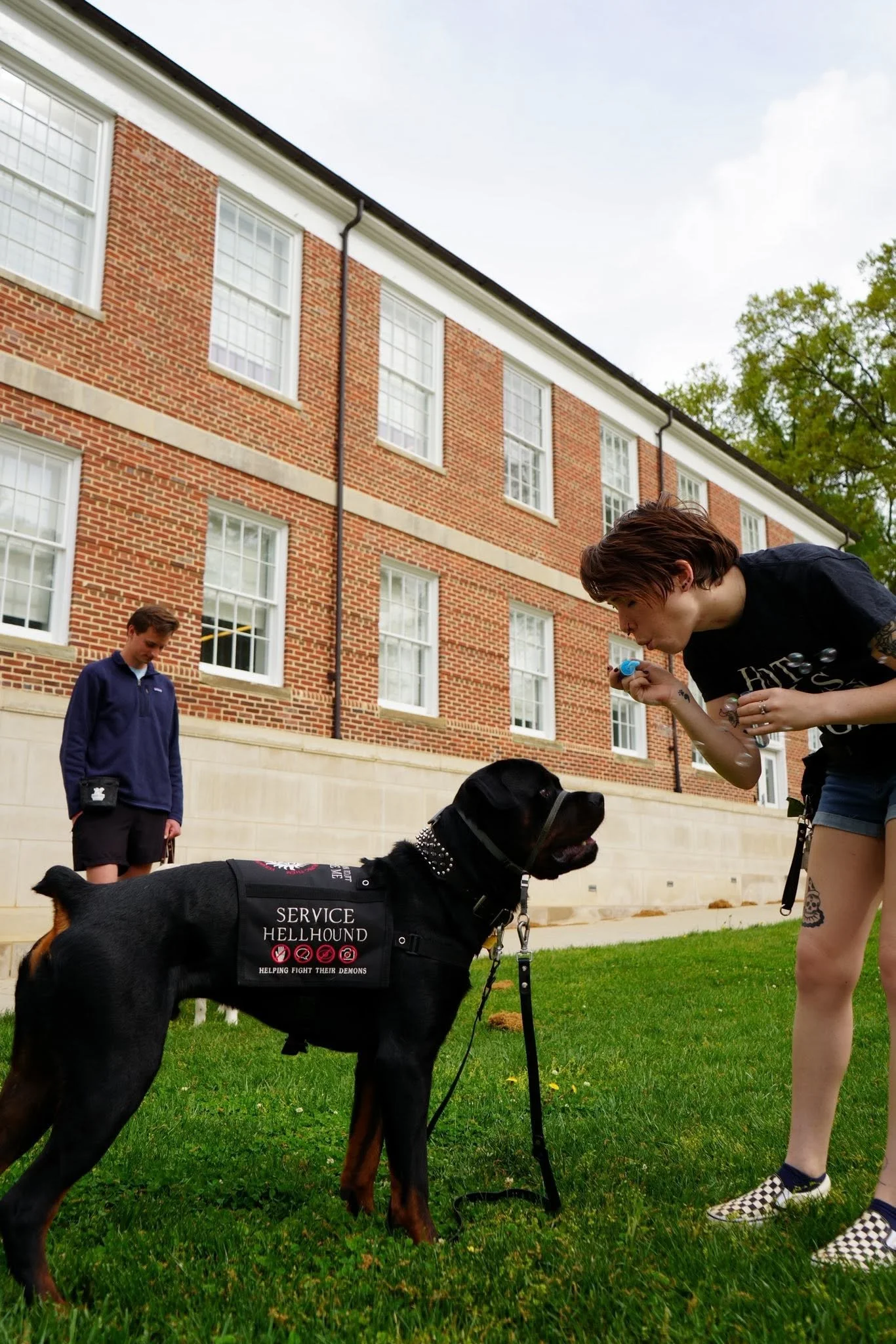 A woman and a service dog, a black and tan Rottweiler mix, in front of a brick building with white-framed windows, green grass, and trees. The service dog is wearing a vest with labels indicating it helps fight demons and assist with service.