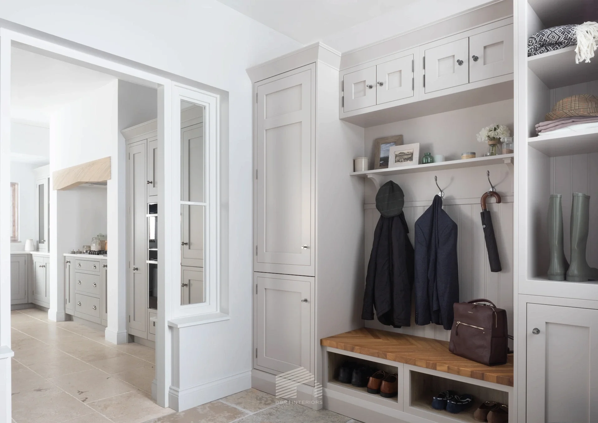 A white entryway with built-in cabinets, a bench, and hooks for hanging coats. There are boots below the bench, handbags, and a few framed pictures and decorative items on a shelf.