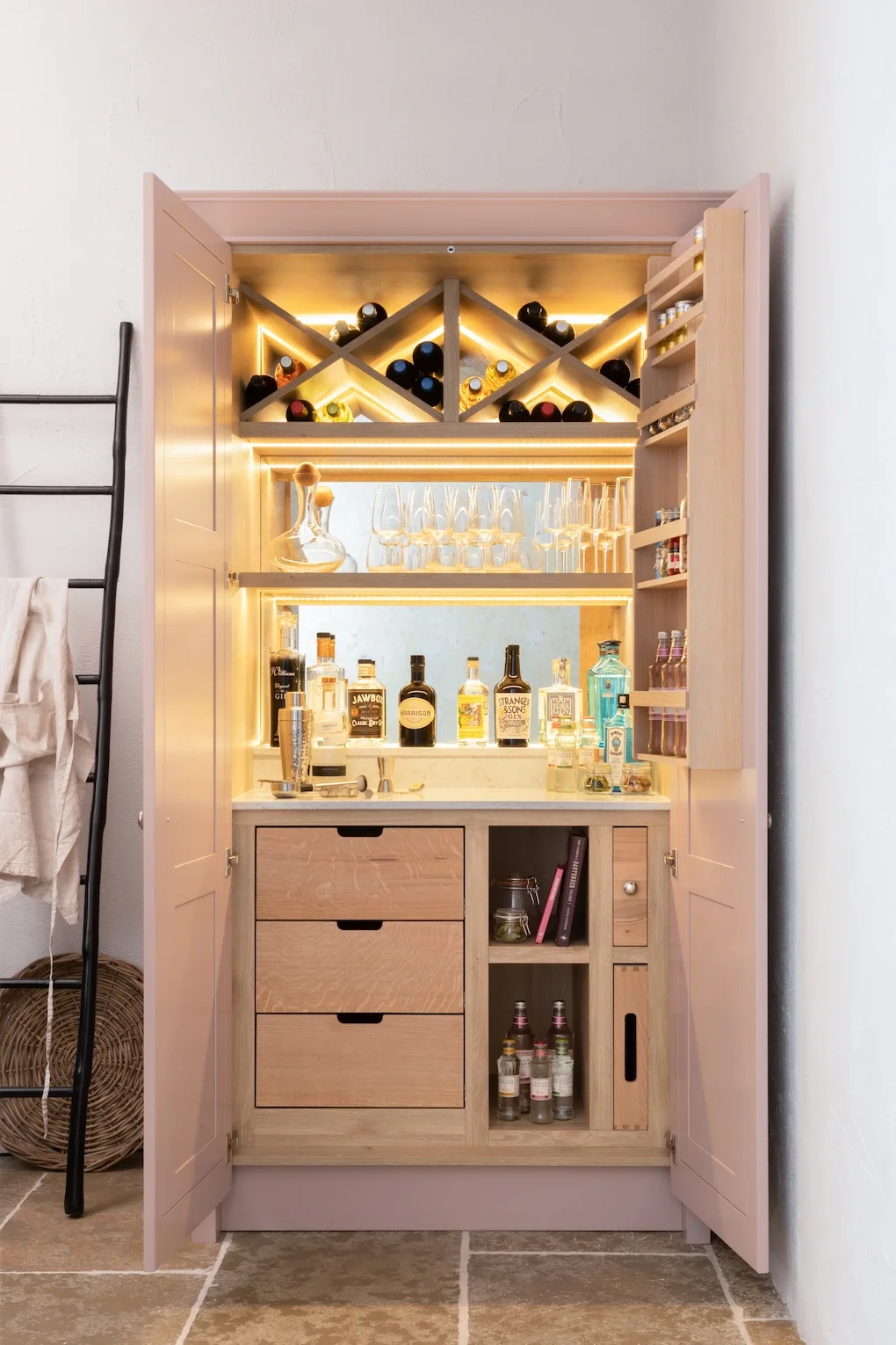 A wooden home bar cabinet with bottles of alcohol, glasses, and a decanter, featuring integrated lighting and wine storage on top.
