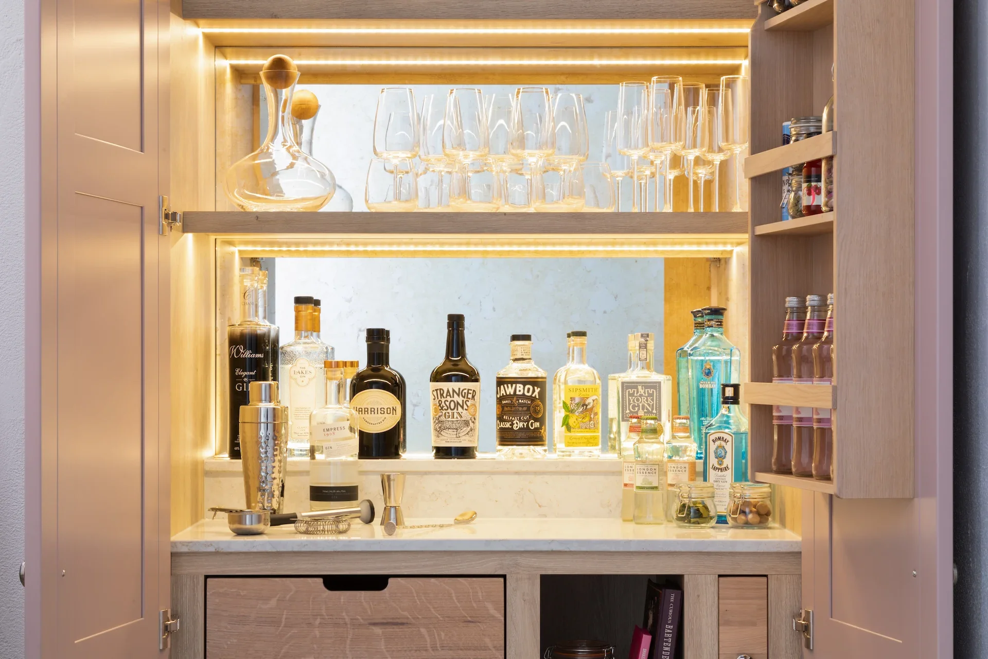 A home liquor bar with illuminated shelves holding various bottles of alcohol, glassware, and bar tools in a wooden cabinet.