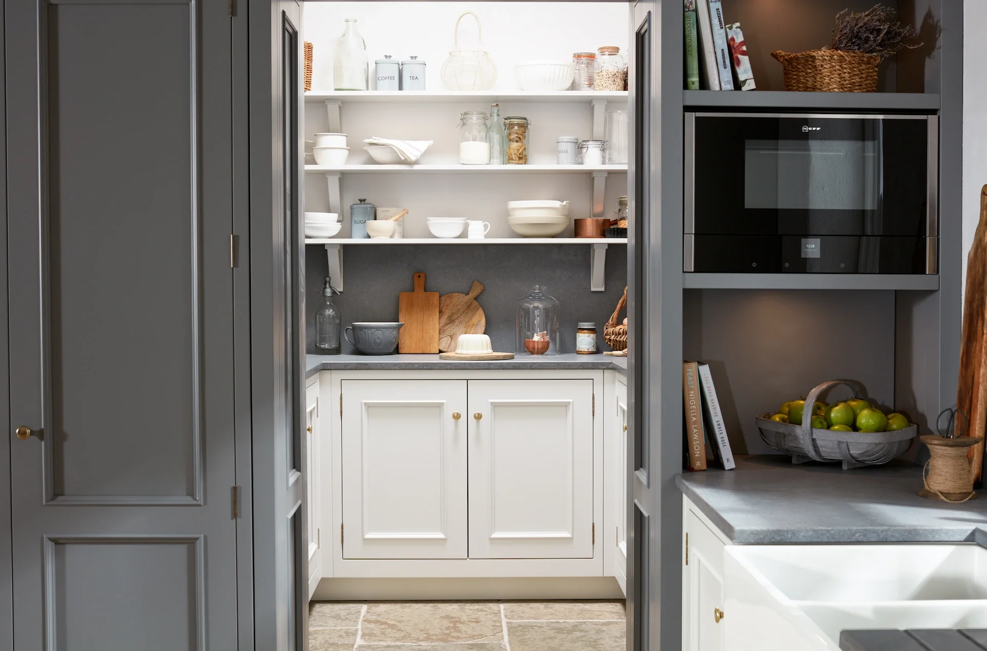View of a kitchen pantry with open shelving, dishes, jars, and kitchenware, seen through a doorway with gray cabinets and a gray countertop.