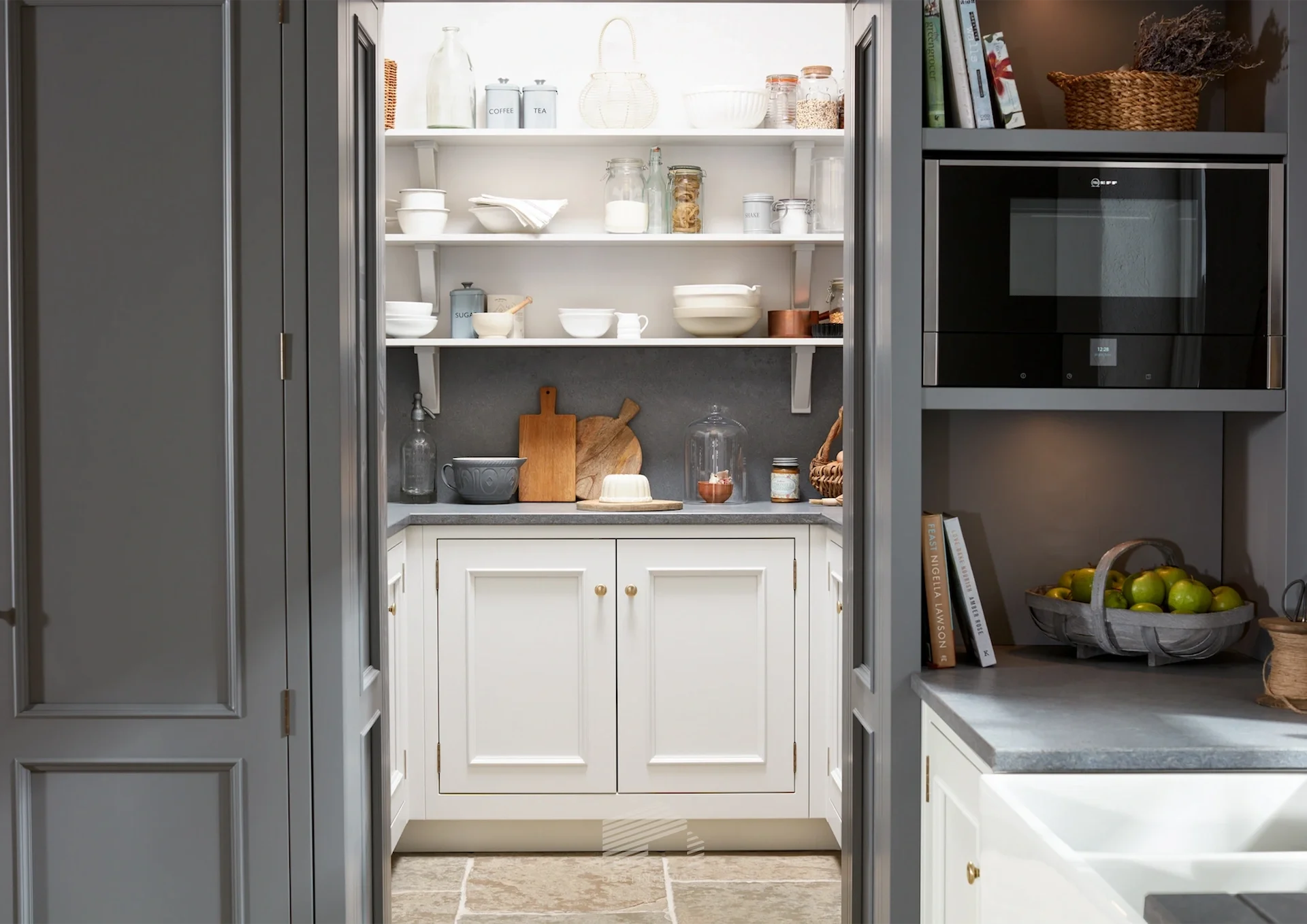 View through an open gray cabinet doorway into a kitchen pantry with shelves holding jars, plates, and a stand mixer.