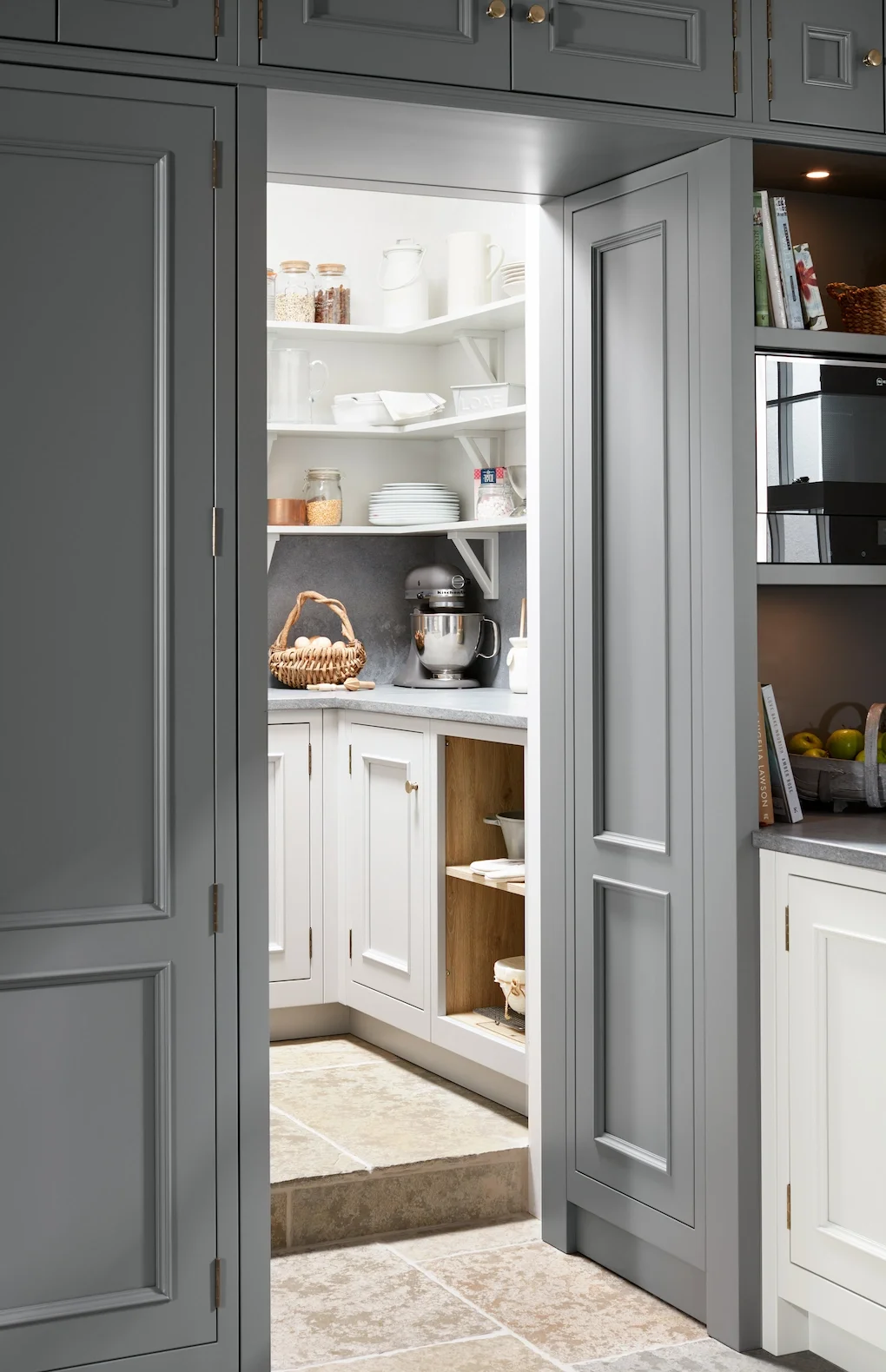 View through an open gray cabinet doorway into a kitchen pantry with shelves holding jars, plates, and a stand mixer.