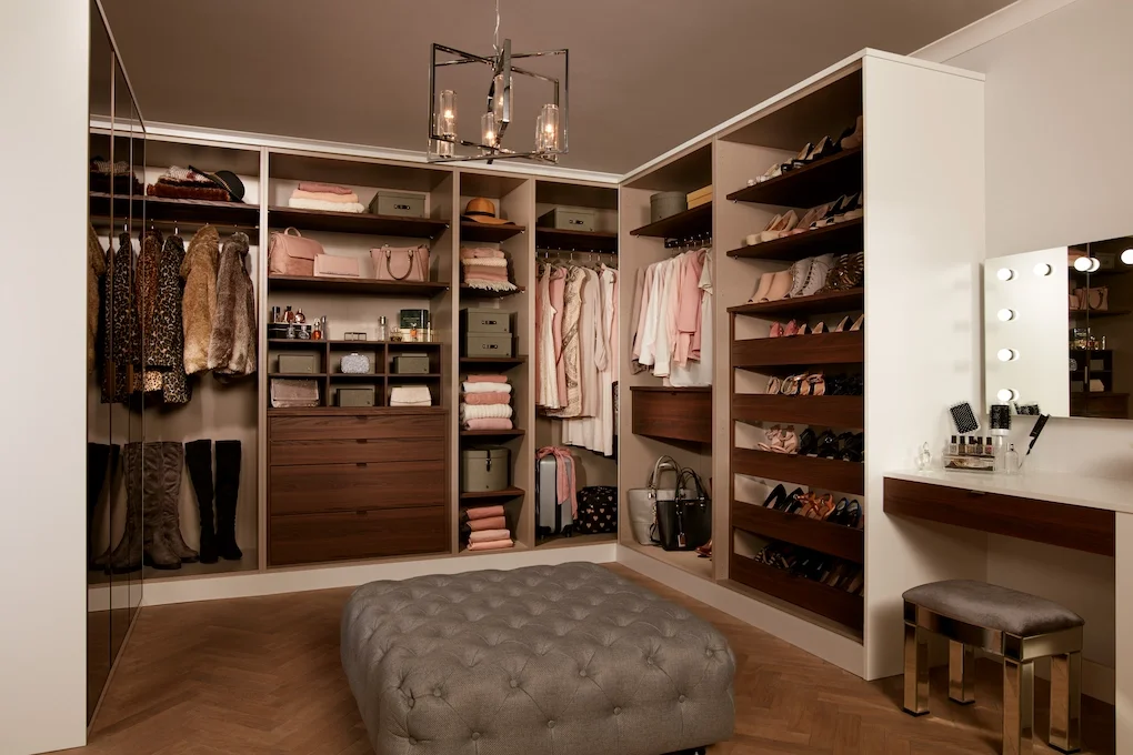 A walk-in closet with clothes, shoes, and accessories organized on shelves and in drawers. A tufted ottoman is in the center, and a vanity with mirror and lights is on the right.
