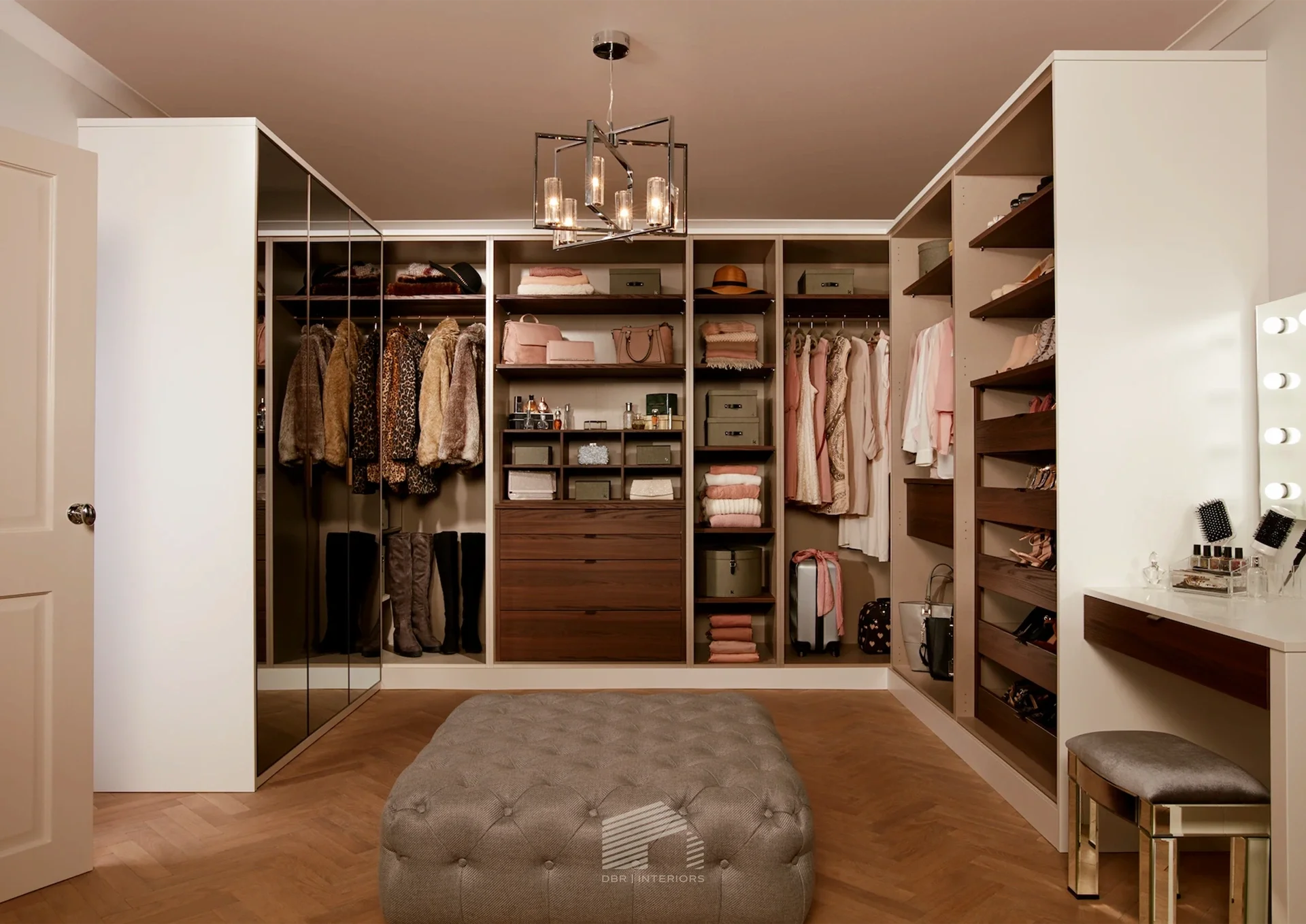 A walk-in closet with clothes, shoes, and accessories organized on shelves and in drawers. A tufted ottoman is in the center, and a vanity with mirror and lights is on the right.
