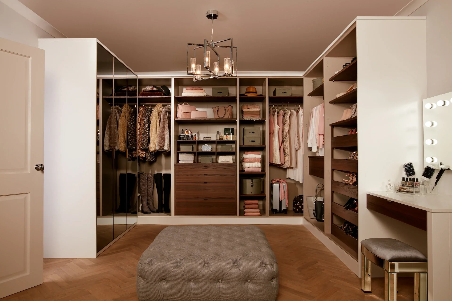 A neatly organized walk-in closet with shelves, hanging space for clothes, and accessories. There's a large tufted ottoman in the middle and a vanity with lights on the right.