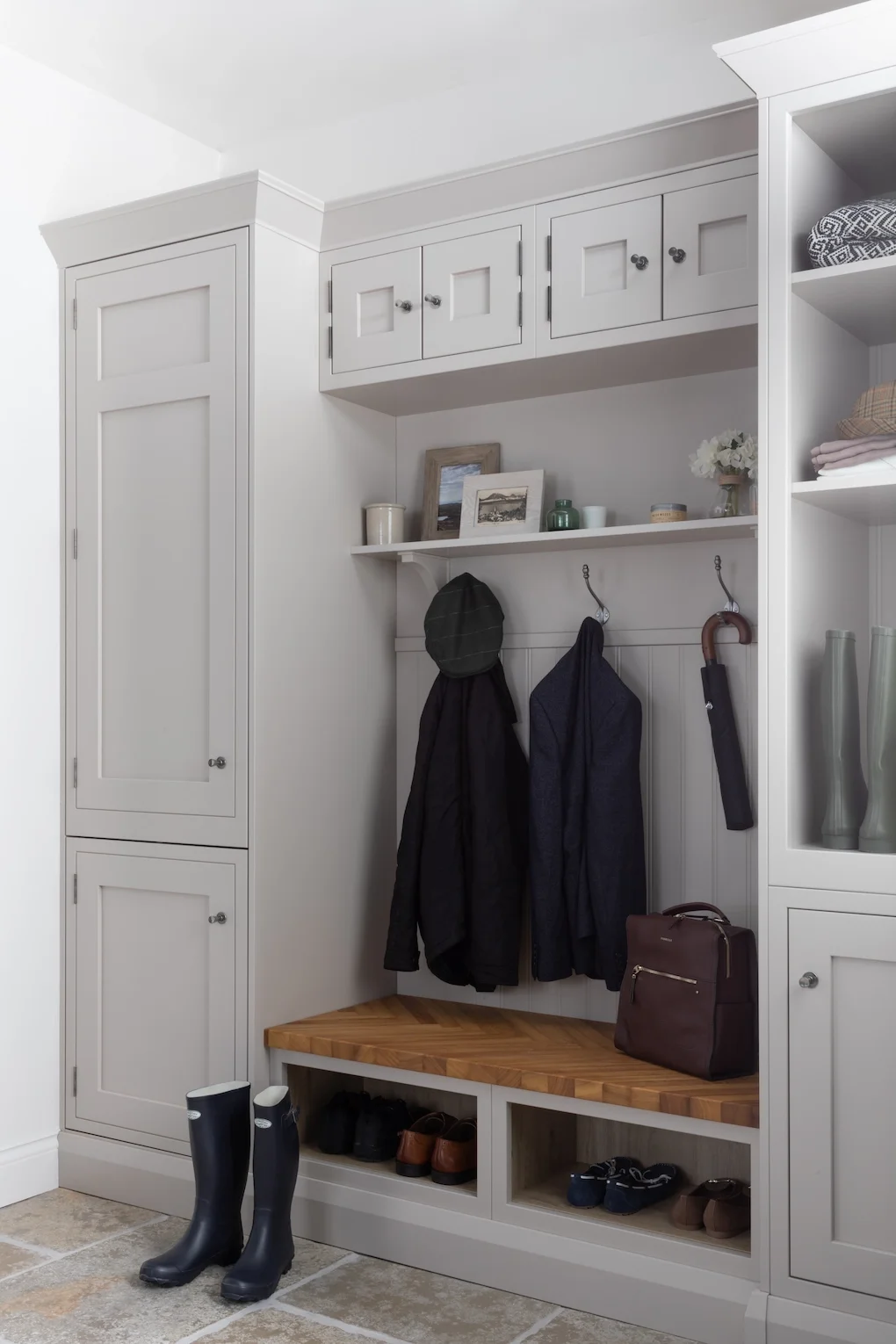 A white entryway with built-in cabinets, a bench, and hooks for hanging coats. There are boots below the bench, handbags, and a few framed pictures and decorative items on a shelf.