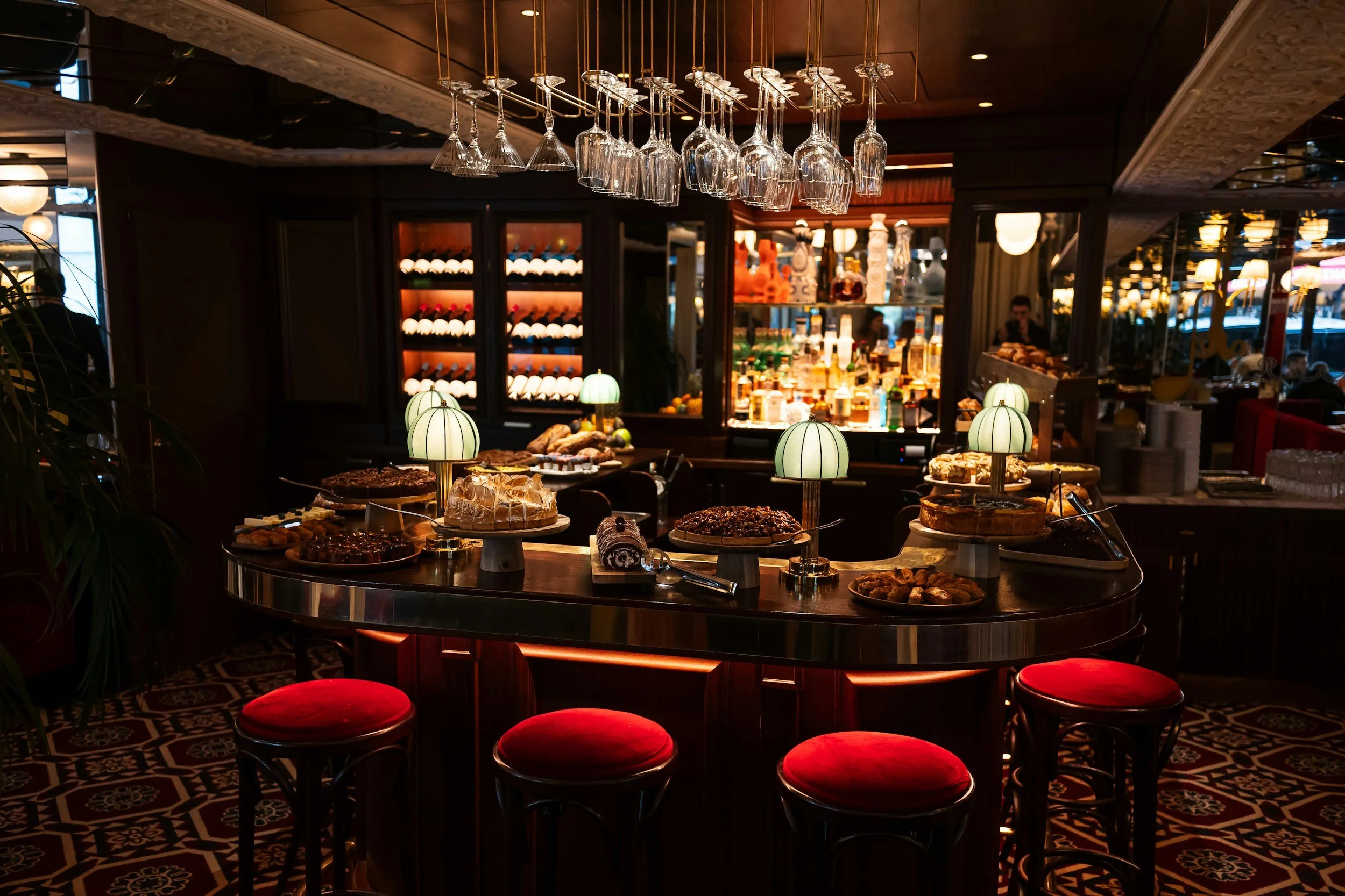 A restaurant buffet with various desserts, cakes, and pastries, illuminated by small table lamps, and with hanging wine glasses above, set against dark wood interior.