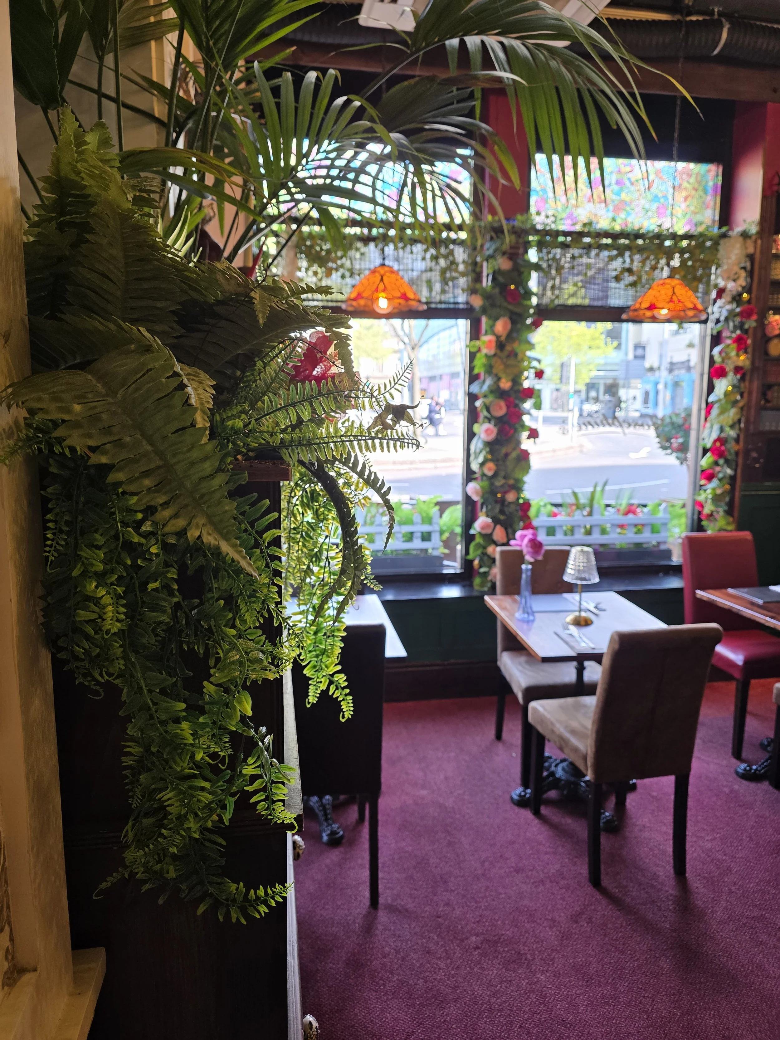 Inside a cozy restaurant with a red carpeted floor, decorated with numerous green plants. There are tables and chairs arranged, some with table lamps and flower centers. Large windows at the back show a city street outside with pedestrians and bikes, with colorful flower decorations around the window area. Hanging lamps emit a warm orange glow.