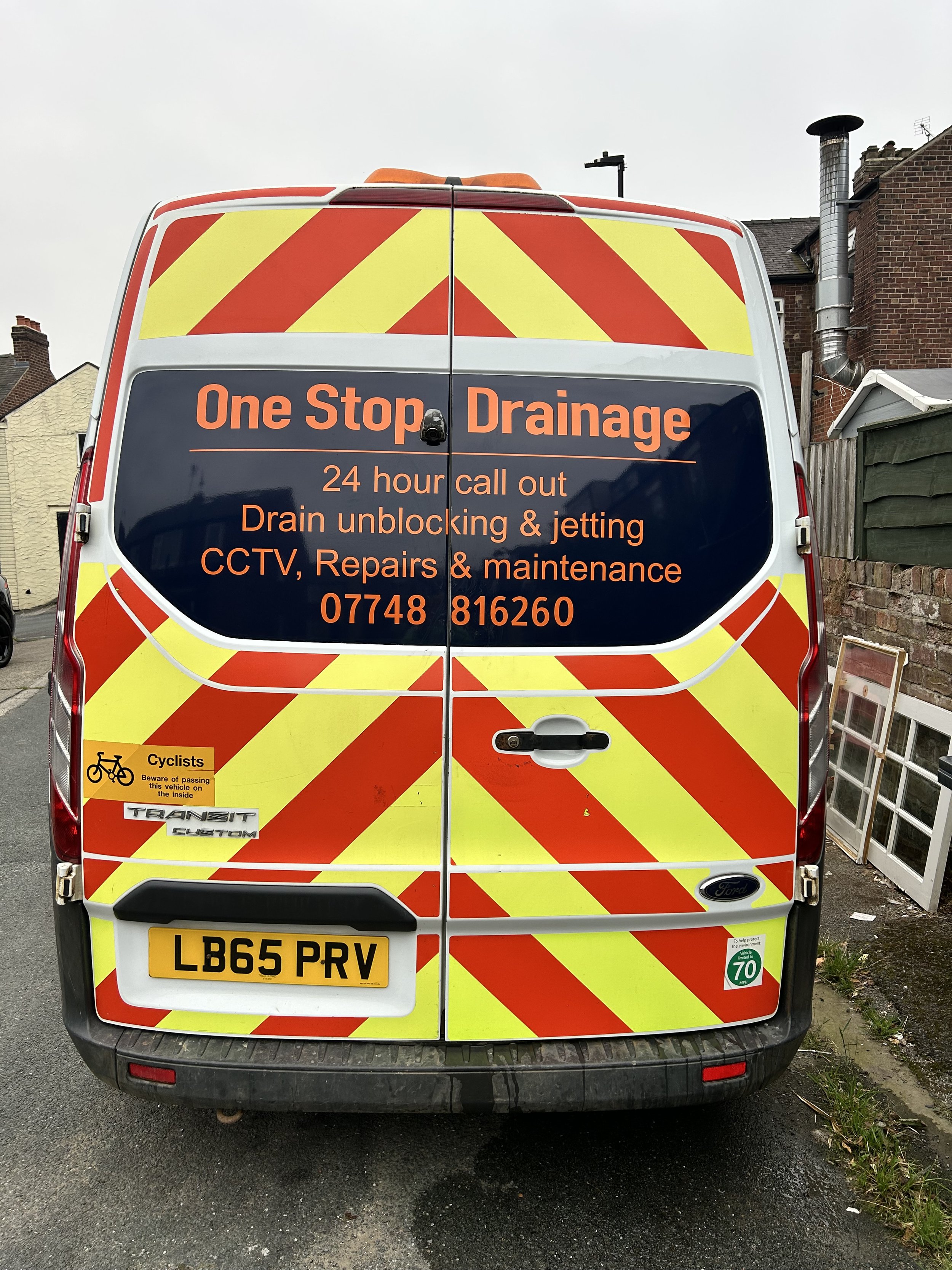 A work van with high-visibility yellow and red chevron stripes on the back, advertising drainage services with contact information, license plate number, and safety stickers.