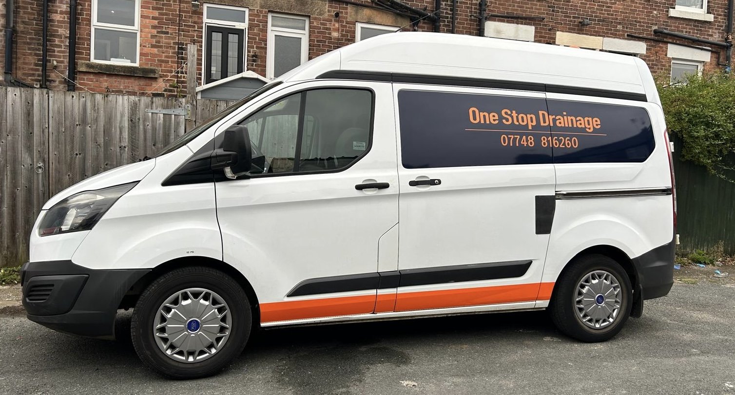 A white service van with orange and black advertising text on the side, parked on a street in front of a wooden fence and brick buildings. The sign reads "One Stop Drainage" with a phone number.