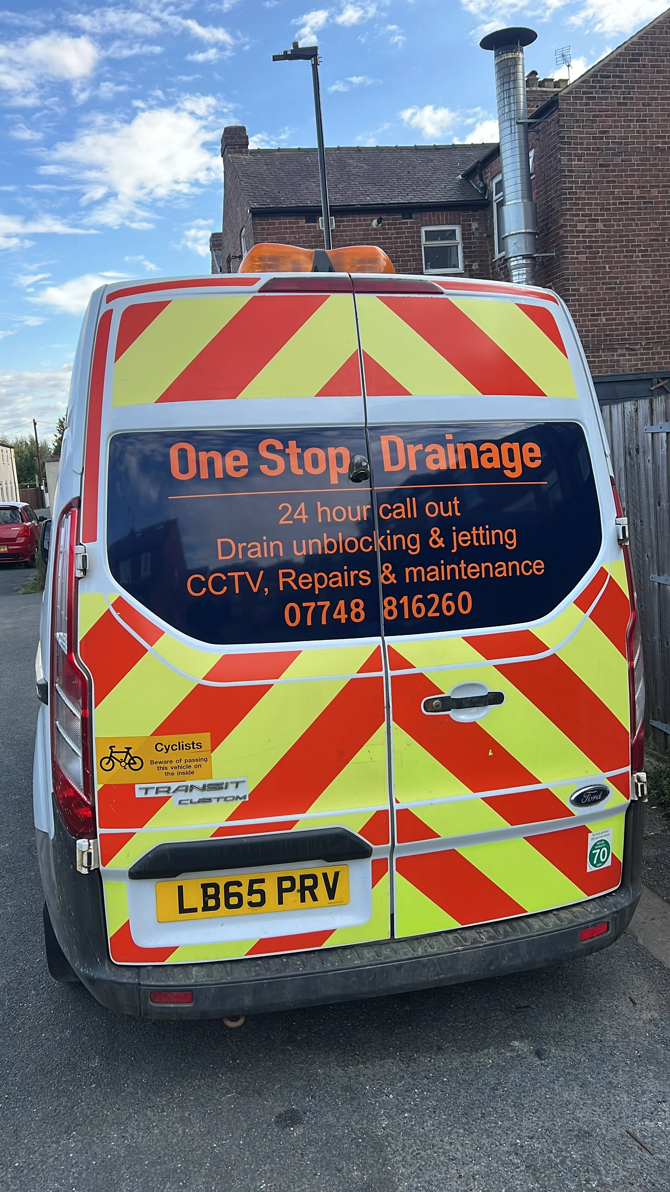 A white service van with yellow and red diagonal stripes on the back doors, advertising drain cleaning and repair services. The sign includes a contact phone number, mentions 24-hour call out, CCTV, repairs, and maintenance. There is a yellow bike lane sign at the bottom left corner.