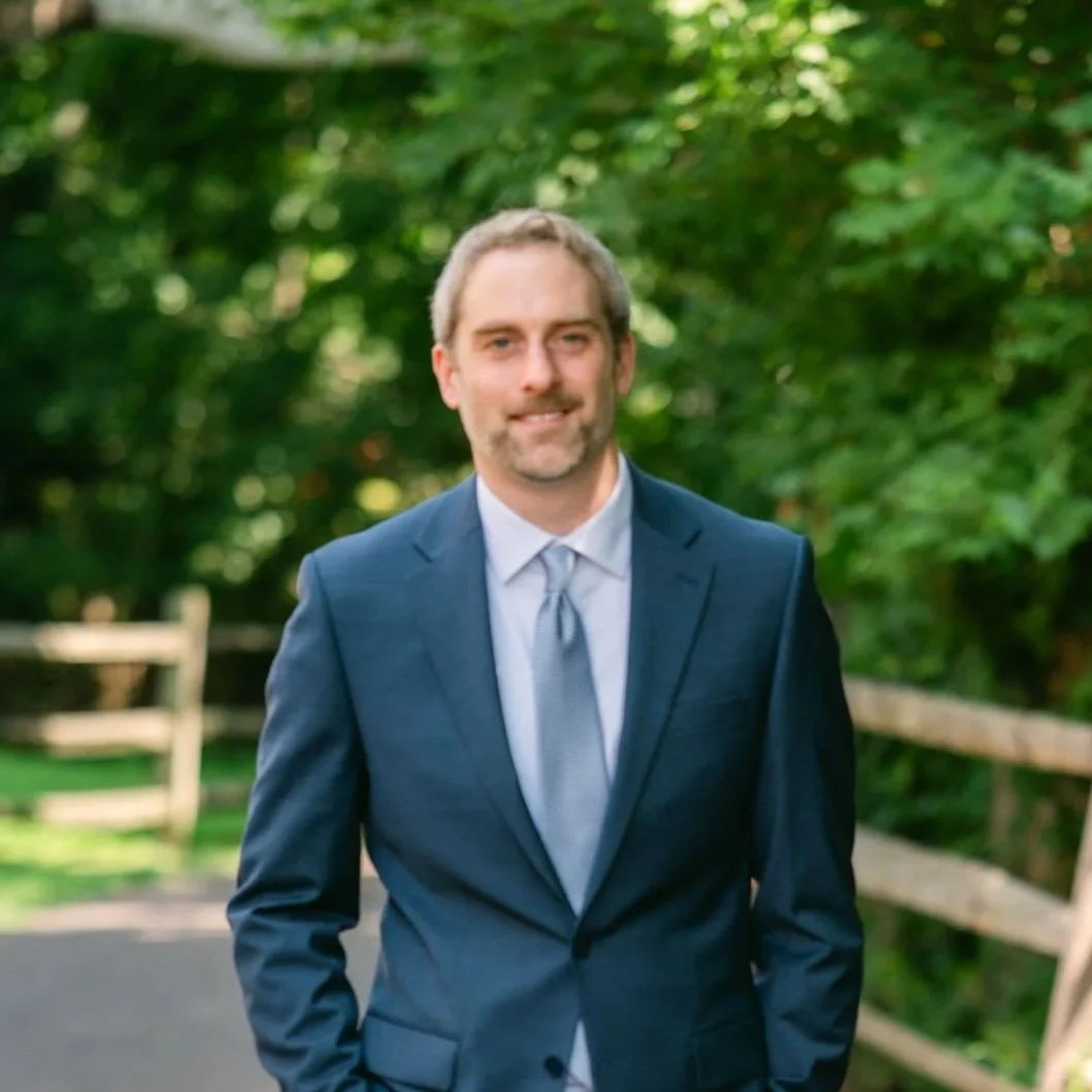 A man wearing a blue suit and light gray tie standing on a paved path with greenery and trees in the background.