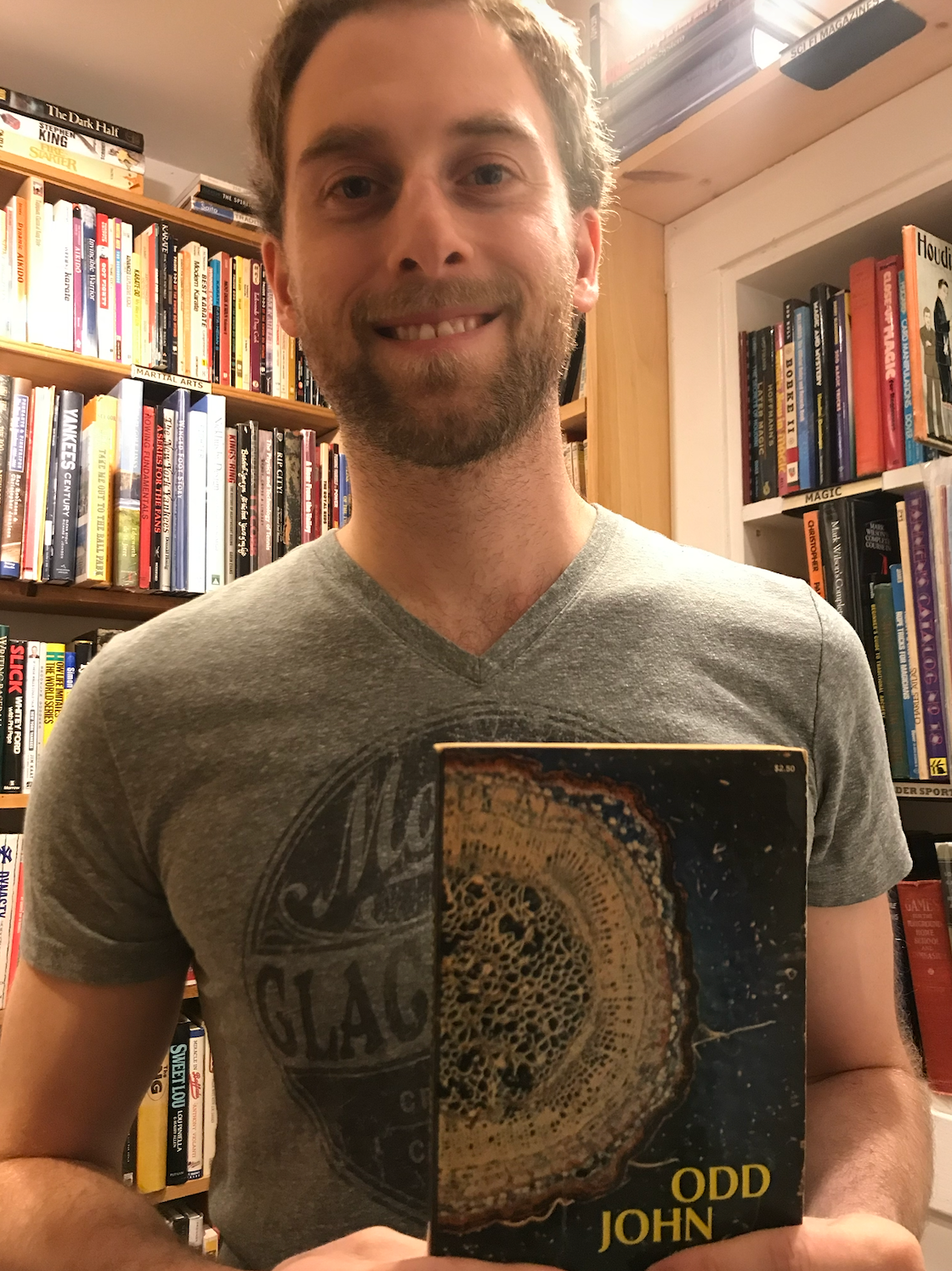 A man with short brown hair and a beard smiling, standing in front of bookshelves filled with books, holding a book titled "Odd John" by Olaf Stapledon in a bookstore or library.