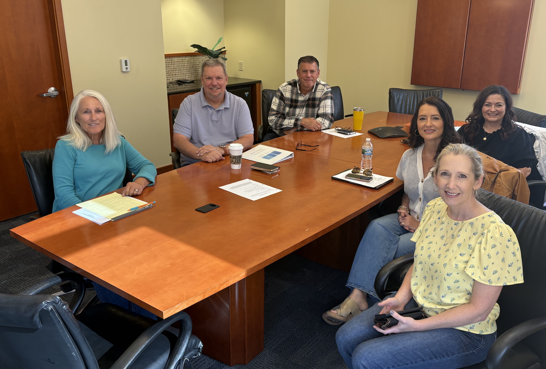 The Trinity Palms Board members sitting around a conference table, smiling at the camera, with notebooks, coffee and cell phones on the table.