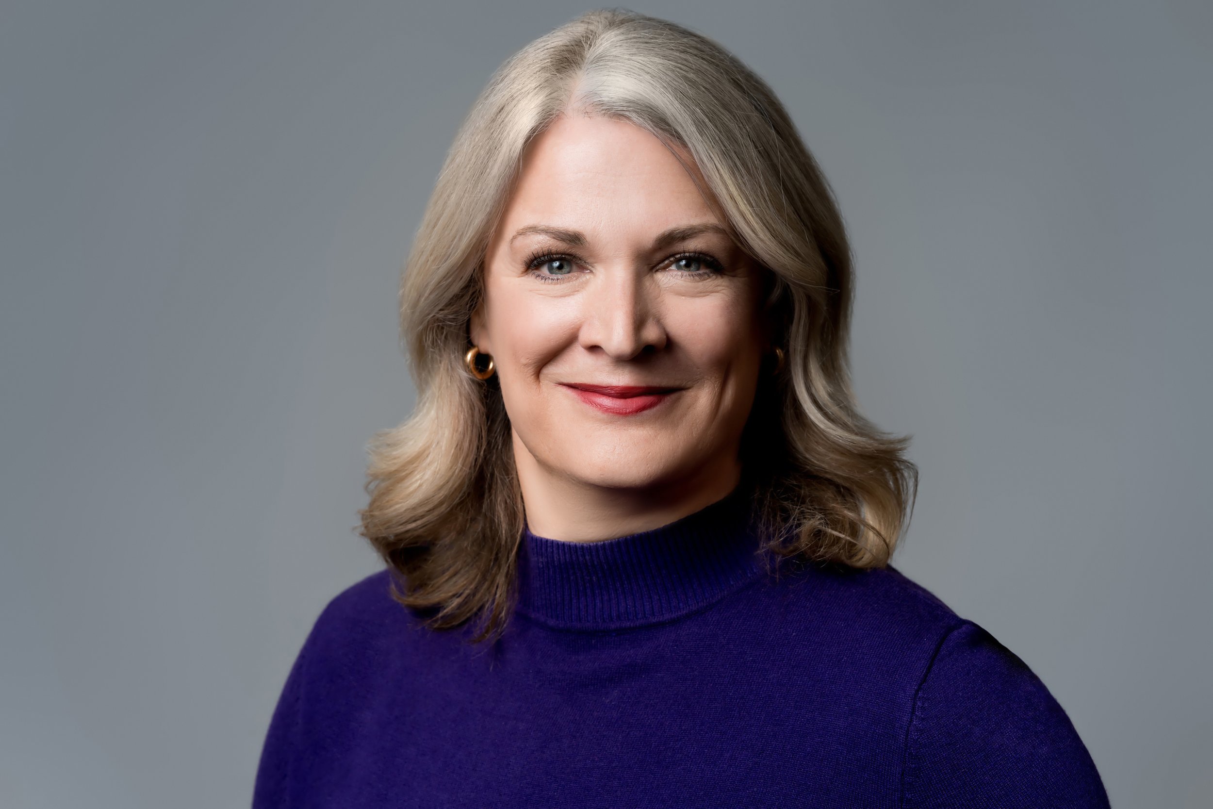 Sharon Ogden, a prayer minister with Trinity Palms, is pictured with shoulder-length gray hair, blue eyes, and wearing a purple sweater, smiling against a gray background.