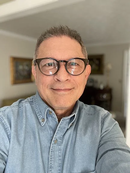 Joshua Gordon, a prayer minister with Trinity Palms Ministries, seen with glasses wearing a light denim shirt, taking a selfie indoors.